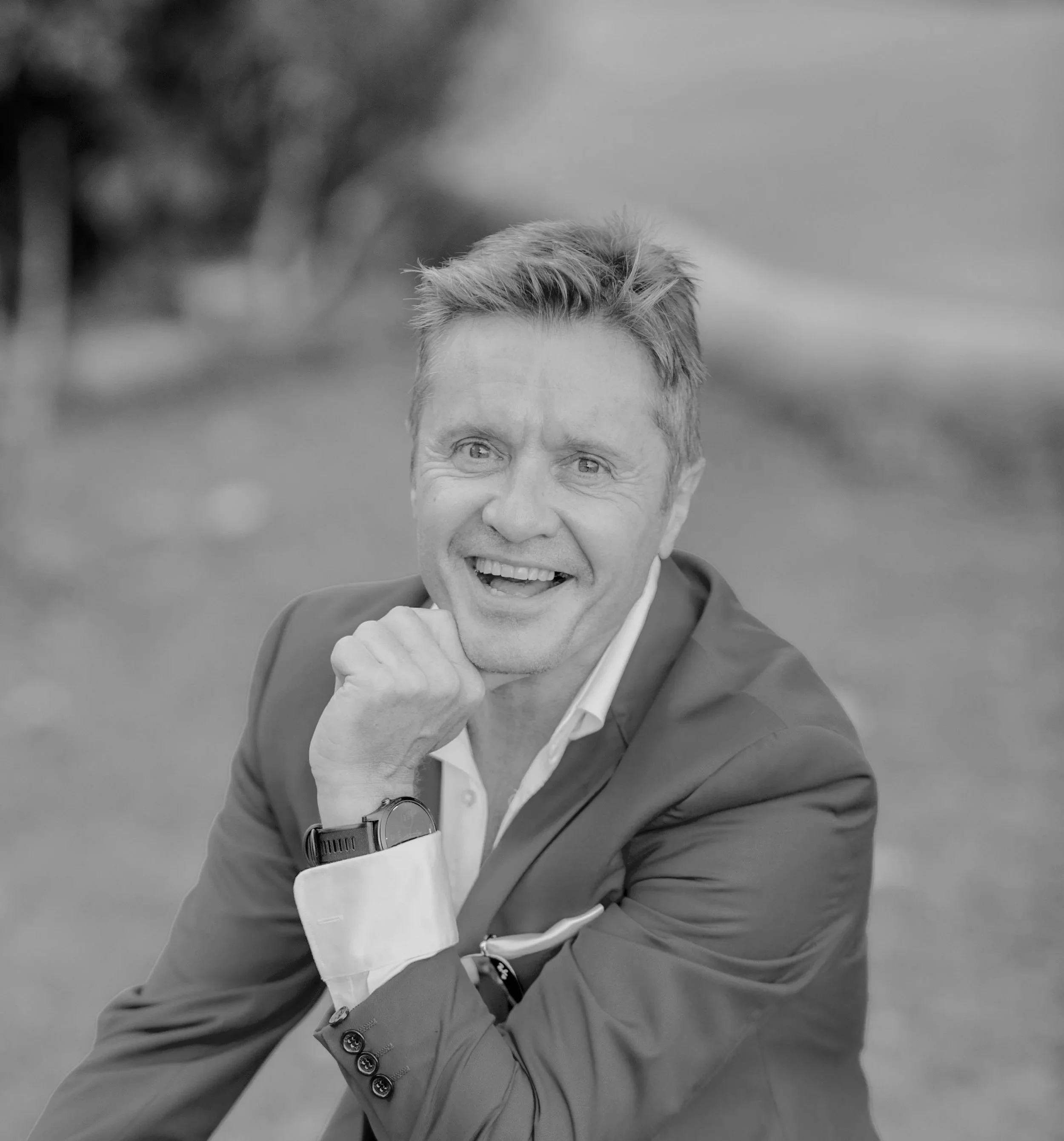 A man smiling outdoors, wearing a suit jacket, with his left hand resting on his chin and looking to the side.