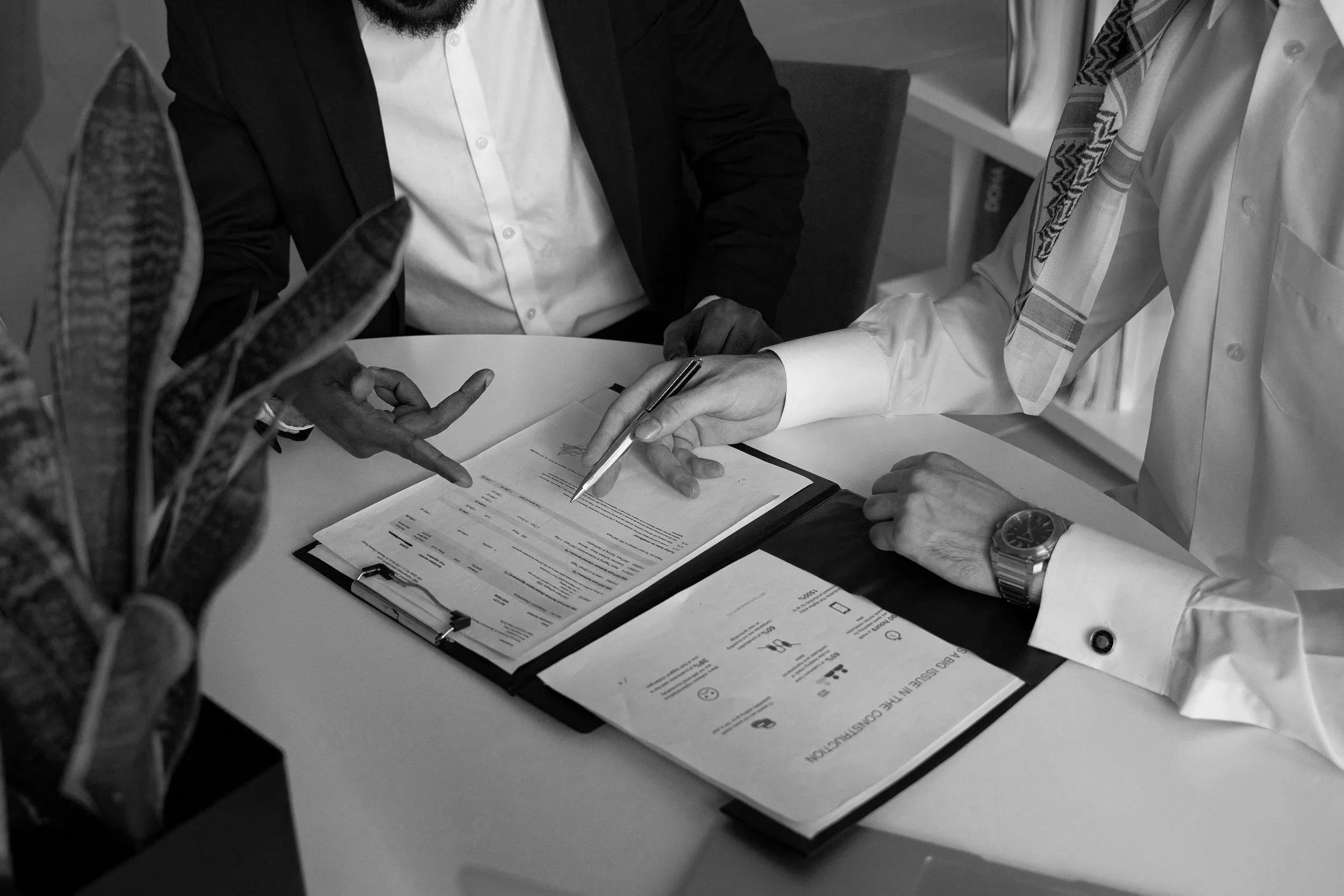 Two men are seated at a table, reviewing documents; one is pointing at the papers, the other is holding a pen.