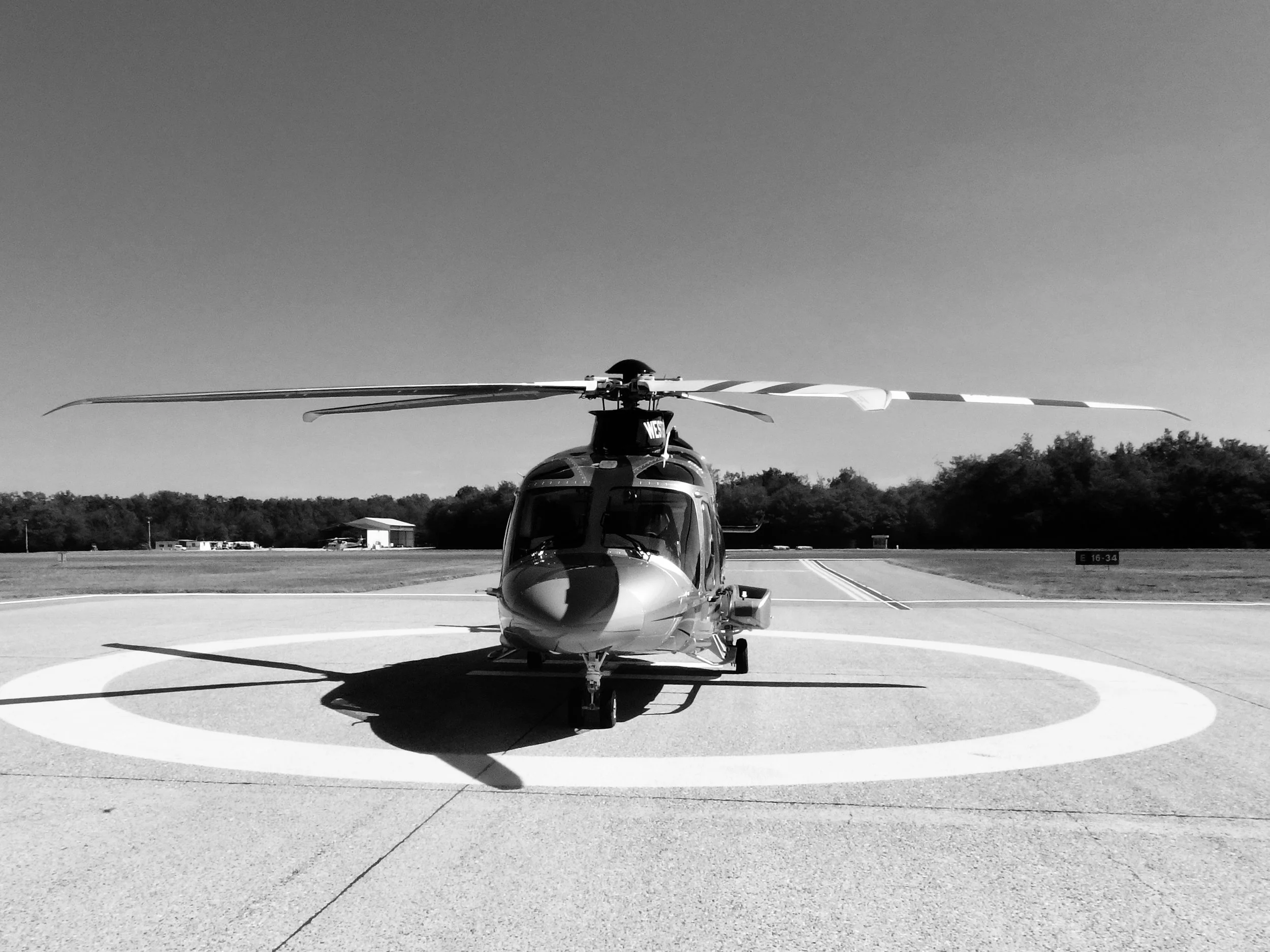 A helicopter parked on a helipad with a circle marking, surrounded by open space and trees in the background, in black and white.