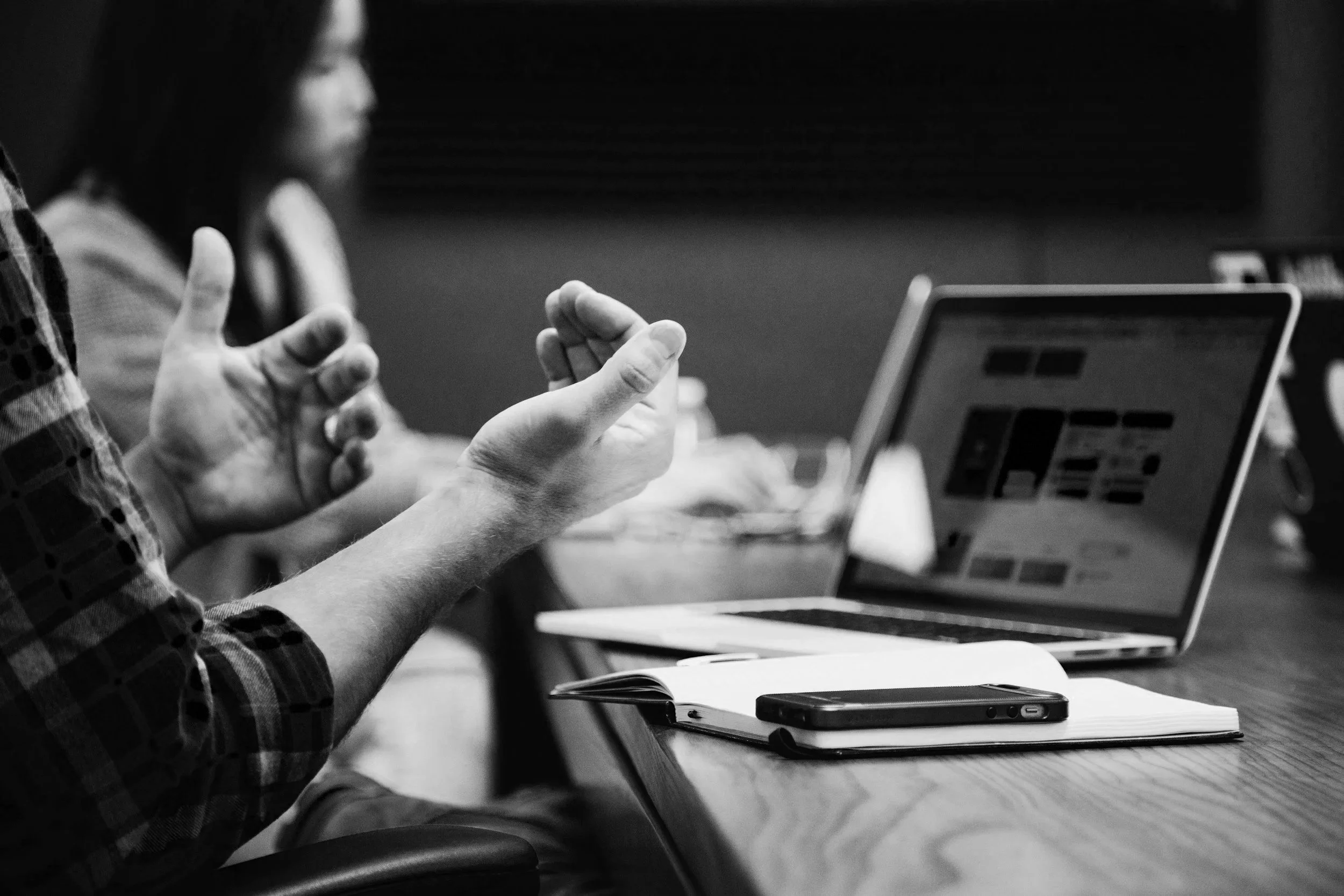 A black and white photo showing two people at a table, one person gesturing with hands while the other looks on. A laptop, a smartphone, and an open notebook are on the table.