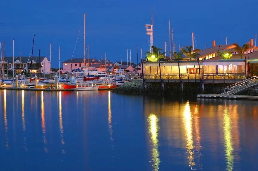 Ben Teakle speaking into a microphone at an outdoor event, wearing sunglasses hanging from his shirt collar and a navy blue polo shirt with a sailboat logo.