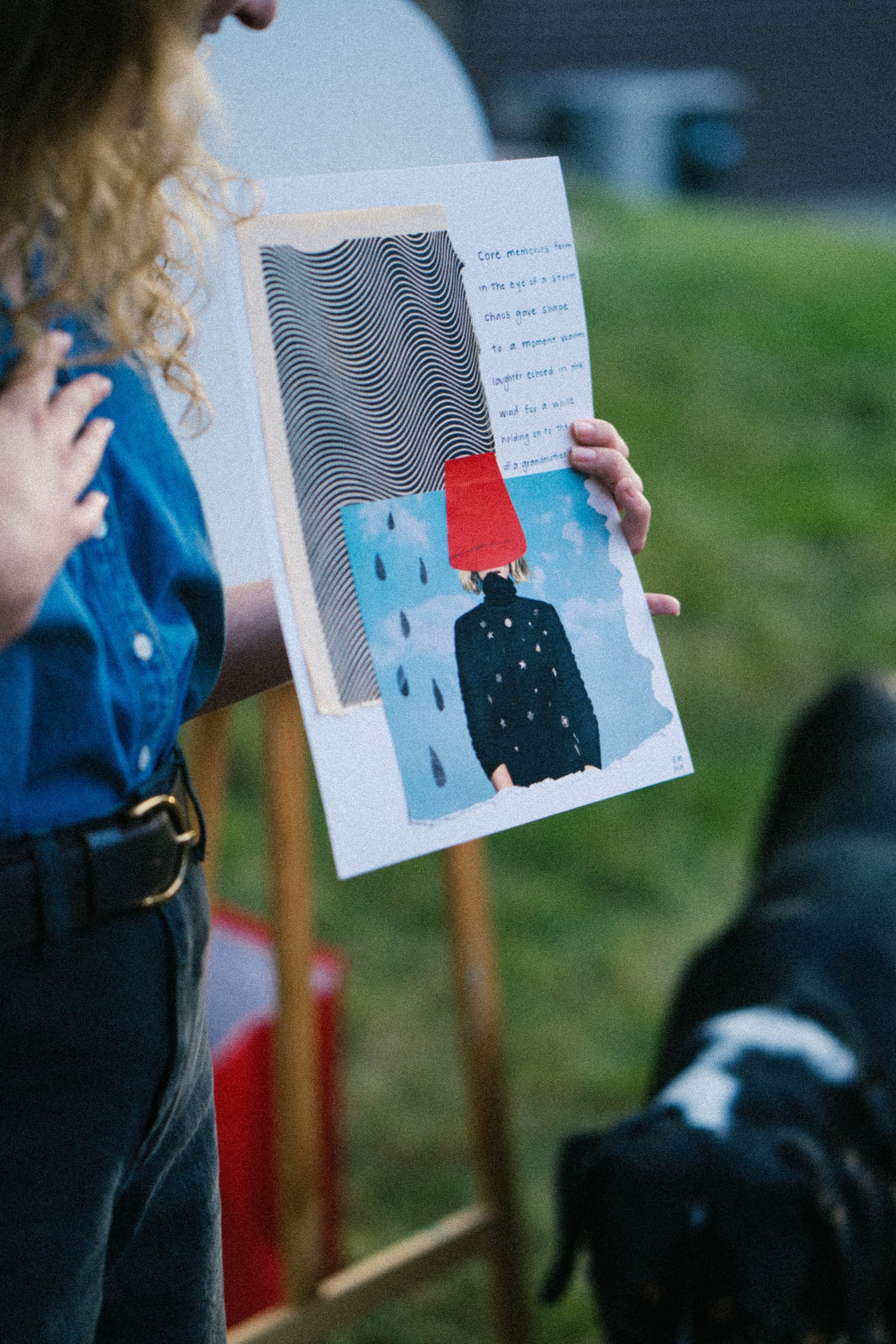 Person holding an art piece featuring a person with a red bucket on their head and a rainy background, with handwritten notes on the side.