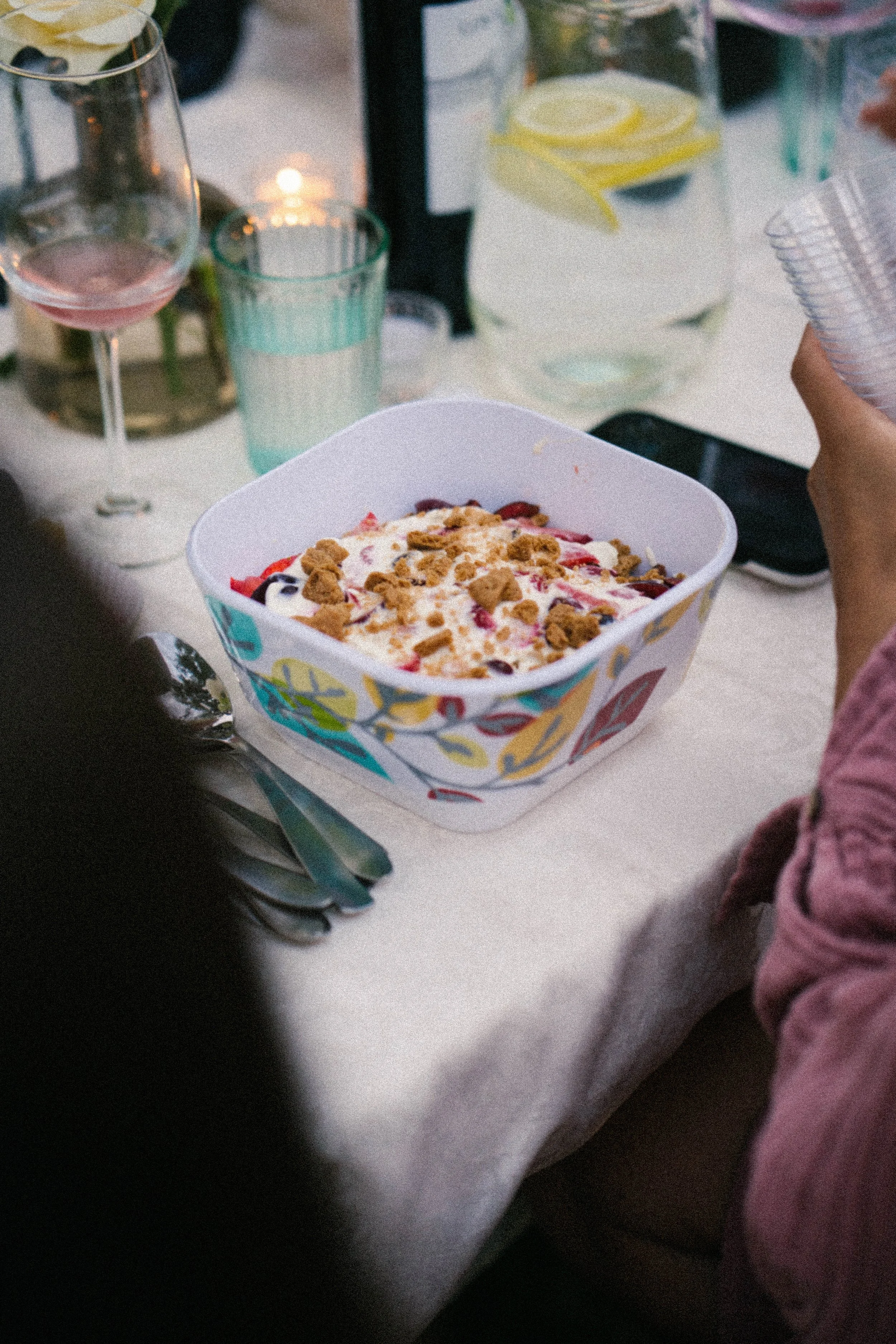 A bowl of fruit salad with granola on top on a dining table with glasses and a pitcher of lemon water.