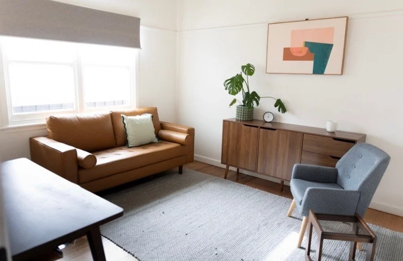 A consulting room with a tan sofa, a light blue armchair, a wooden sideboard with a plant, a clock, and a white mug, a large window with a gray blind, a piece of abstract wall art, and a light gray area rug.