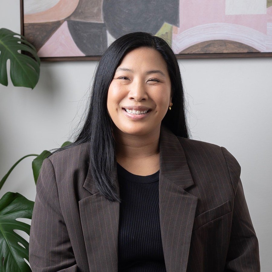 A woman with long black hair smiling, wearing a pinstripe blazer and a black top, sitting in front of a white wall with abstract artwork and green plants.
