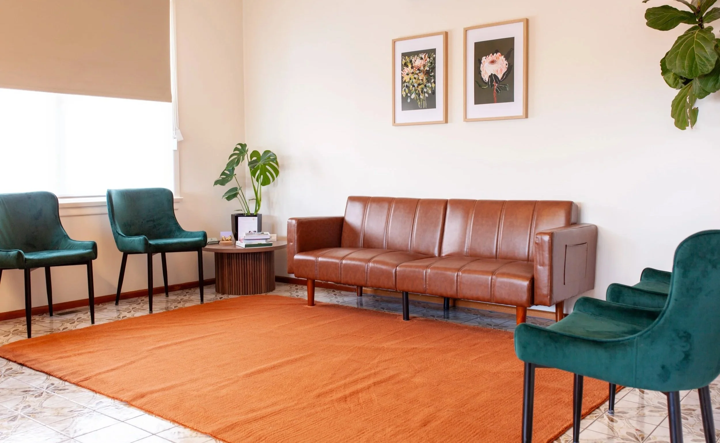 A cozy waiting room with a brown leather sofa, green velvet armchairs, a small round wooden table with books, a potted plant, and wall art of floral prints.