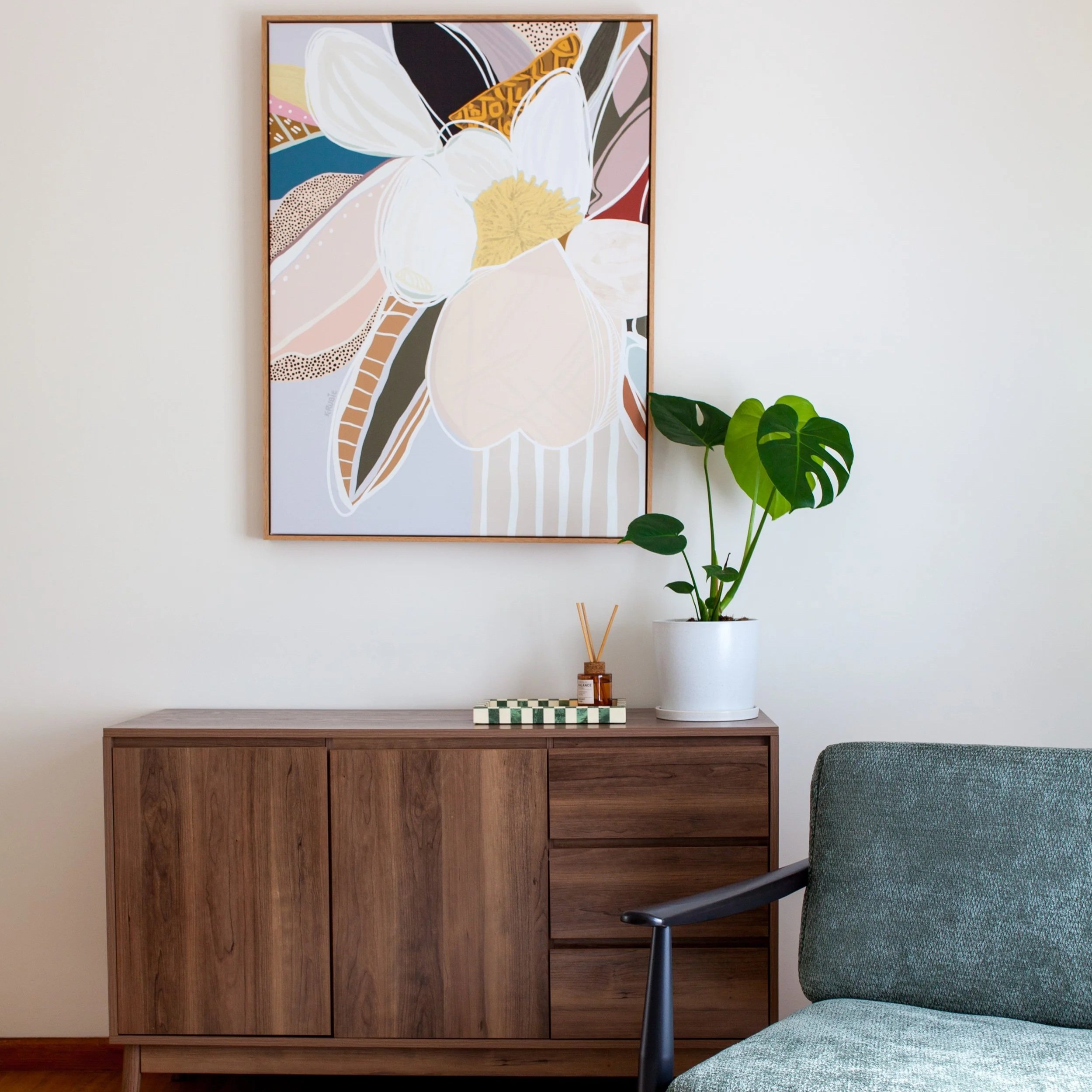Interior of a room featuring a wooden mid-century modern cabinet, a framed abstract floral artwork, a potted plant with broad green leaves, a geometric-patterned tray with a reed diffuser, and a green upholstered chair.