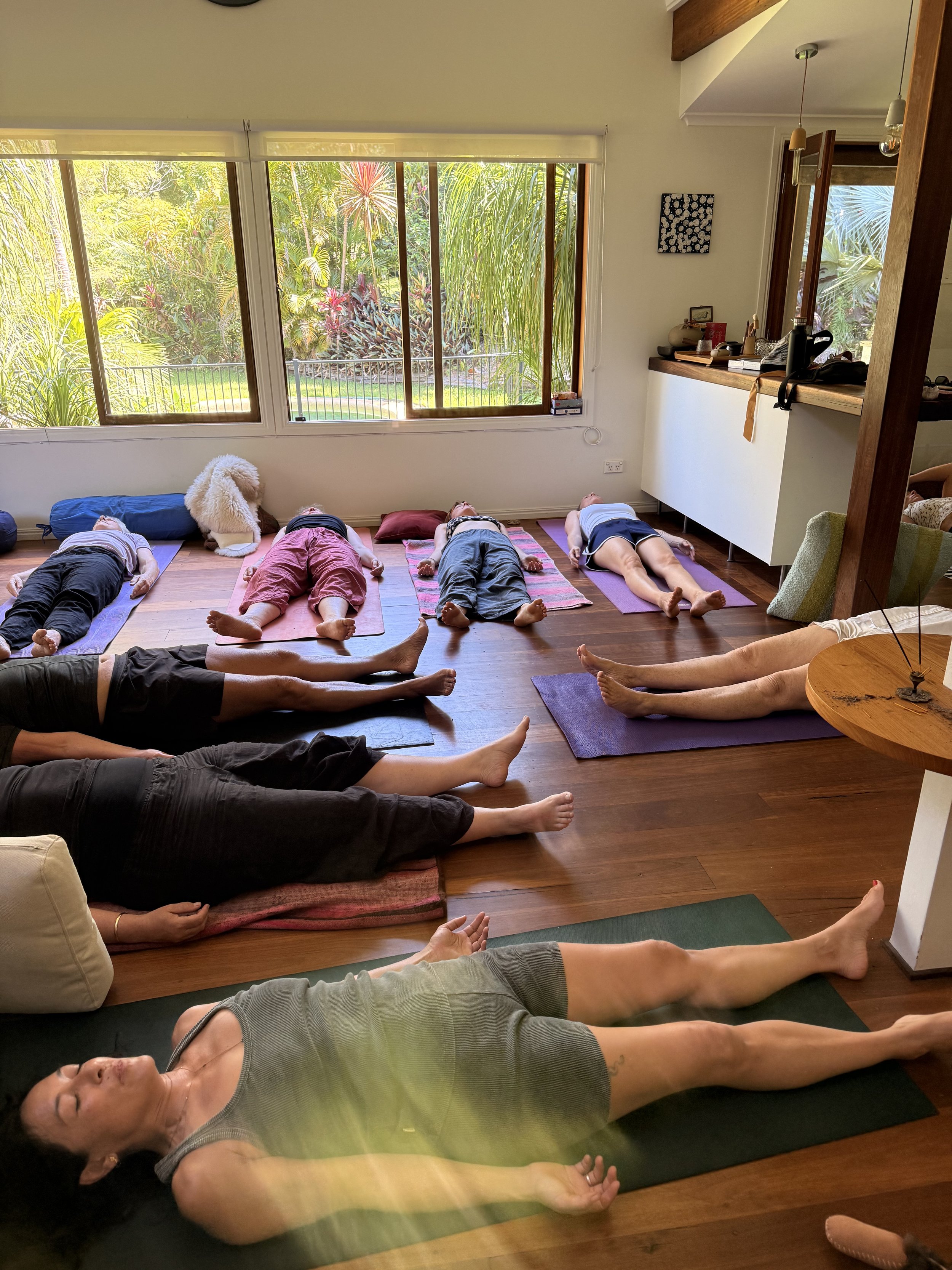 People lying on yoga mats in a bright room with large windows overlooking a garden during a yoga class.