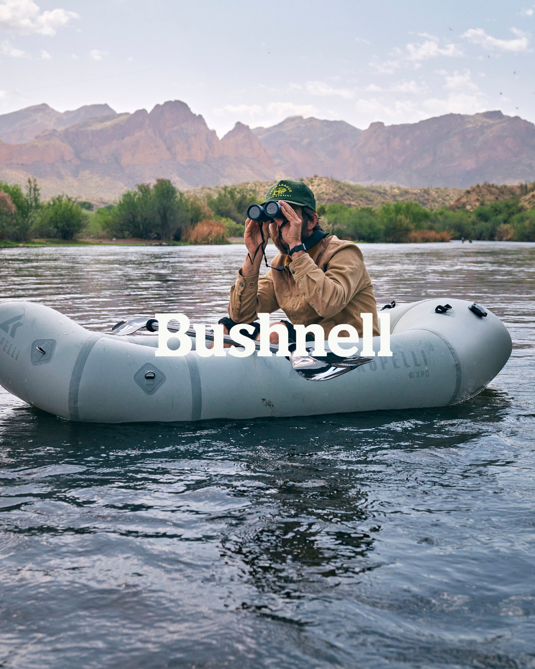 A person in a brown jacket using binoculars while sitting in an inflatable boat on a river with mountains and green trees in the background.