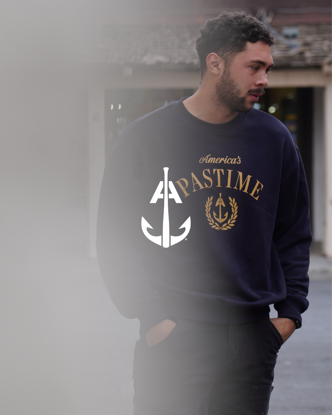 A man with dark, curly hair and a beard wearing a navy sweatshirt with the words "America's Pastime" and a logo featuring a nautical anchor on the front, standing outdoors with a blurred background.