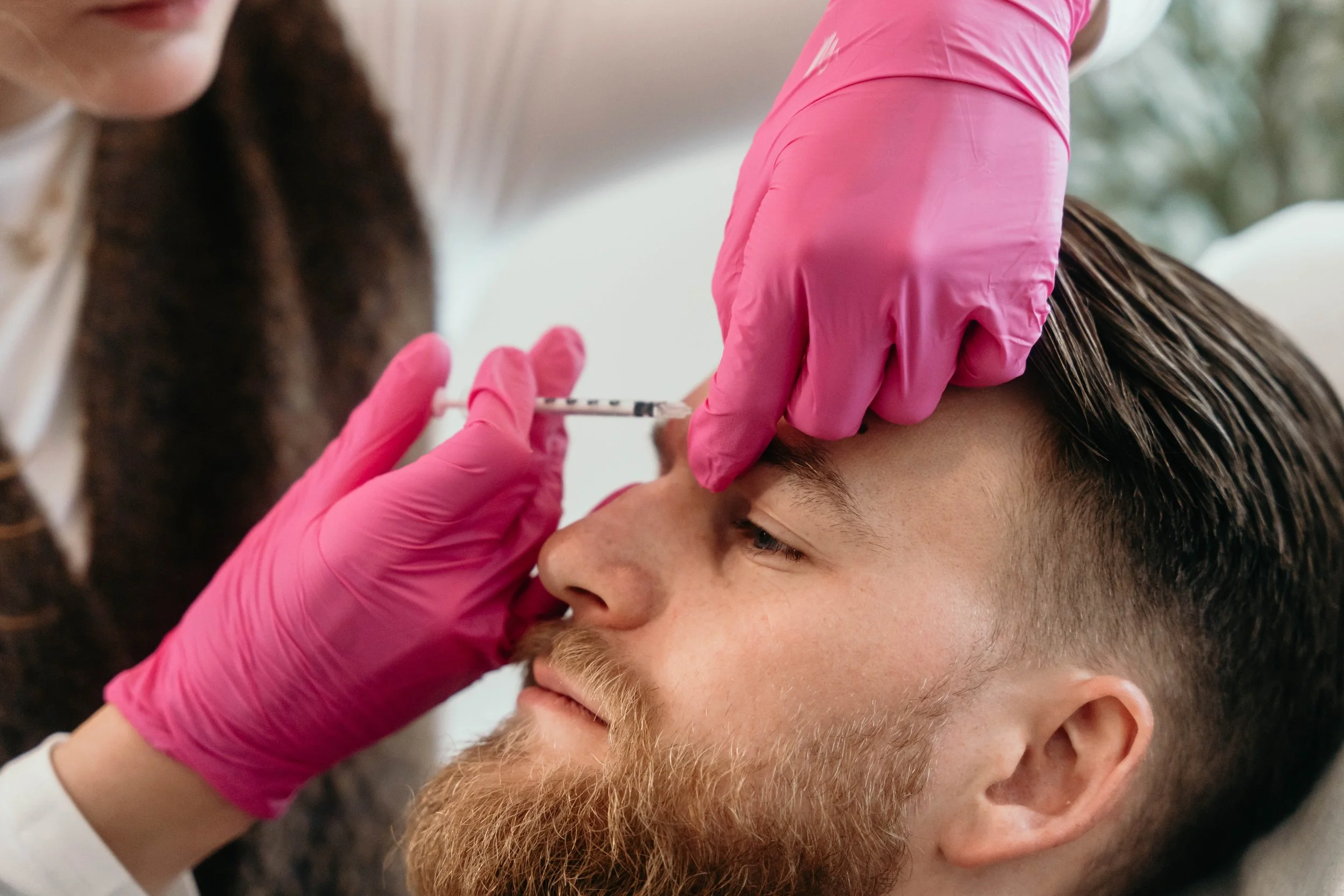 A person receiving a cosmetic injection in the forehead, administered by another person wearing pink gloves.