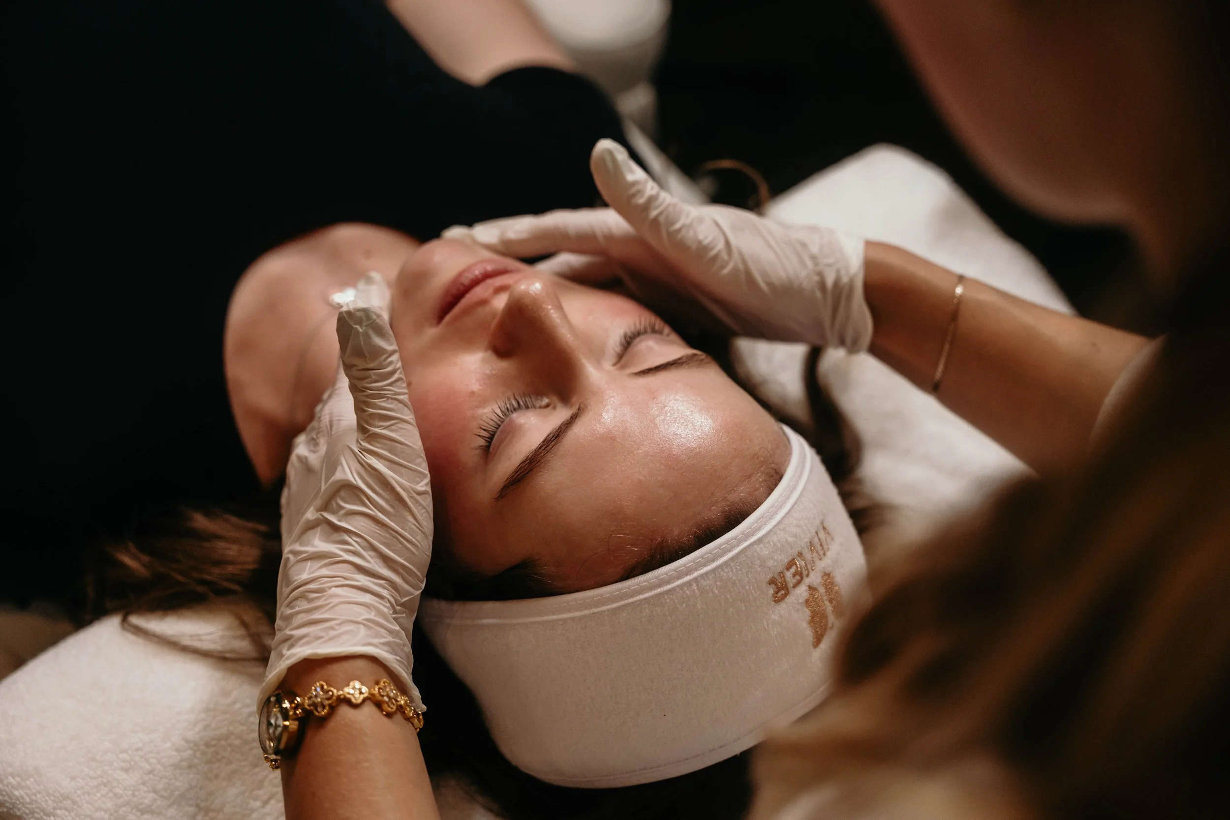 A person receiving a facial massage at a spa, wearing a headband and attended by someone with gloves.