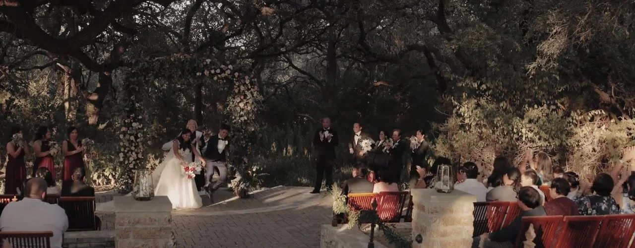 Wenjane and Susie walking the aisle as guests clap under the Sacred Oaks