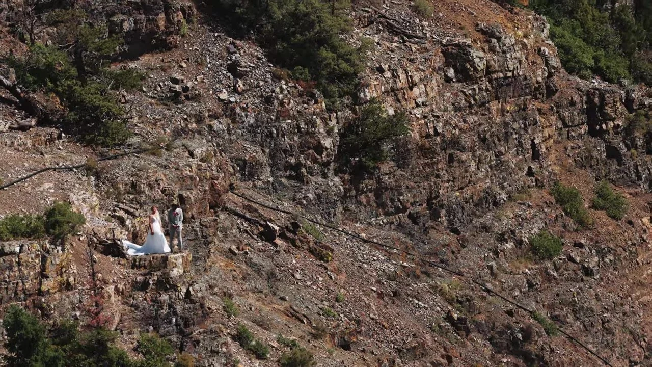 Wide aerial of the couple on a rocky ledge against a huge cliff face at Gold Mountain, Ouray