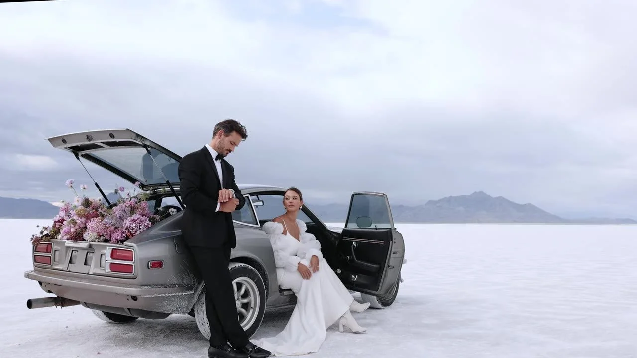 Wide shot of the couple at the vintage 280Z on the salt flats, groom checking his watch, hatch filled with flowers