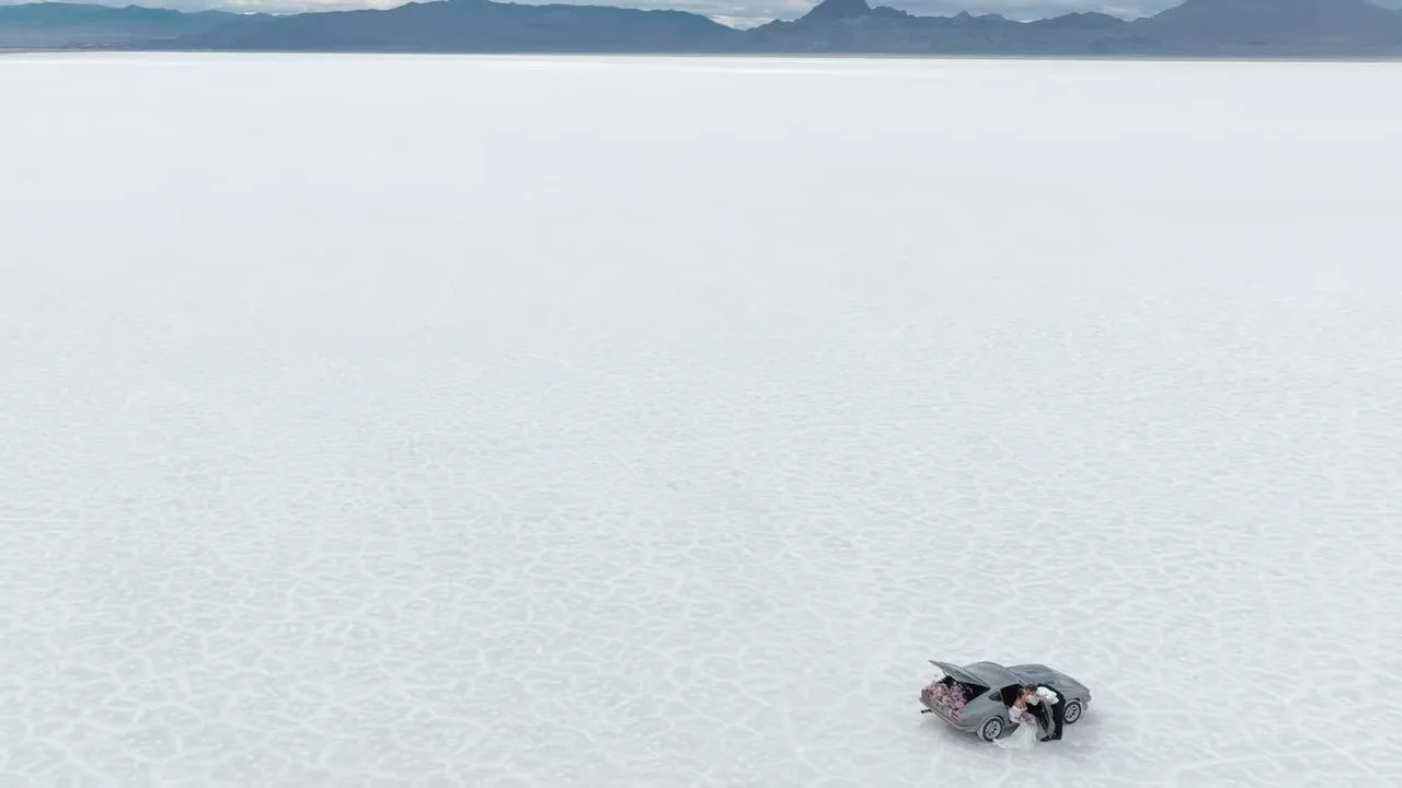 High aerial of the couple and vintage Datsun 280Z on the cracked Bonneville Salt Flats, hatch filled with pink flowers