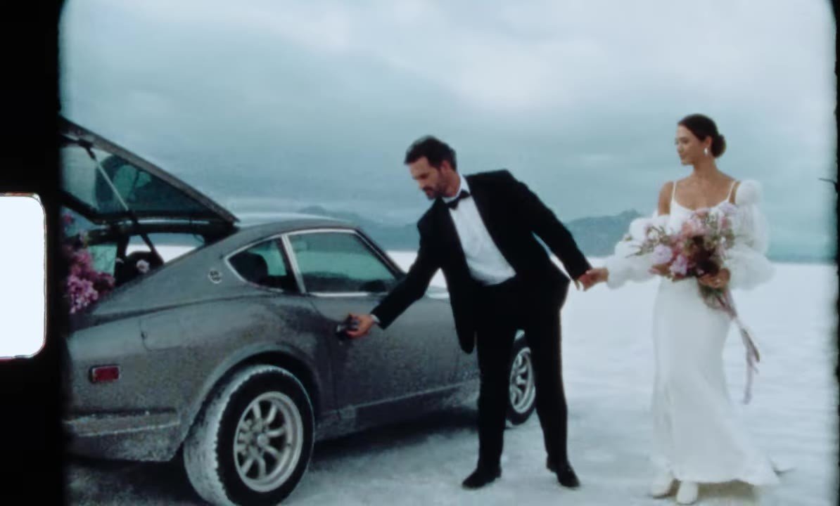 Super 8 frame of the couple at the open door of a vintage 280Z on the salt flats, hatch filled with pink flowers, sprocket edge