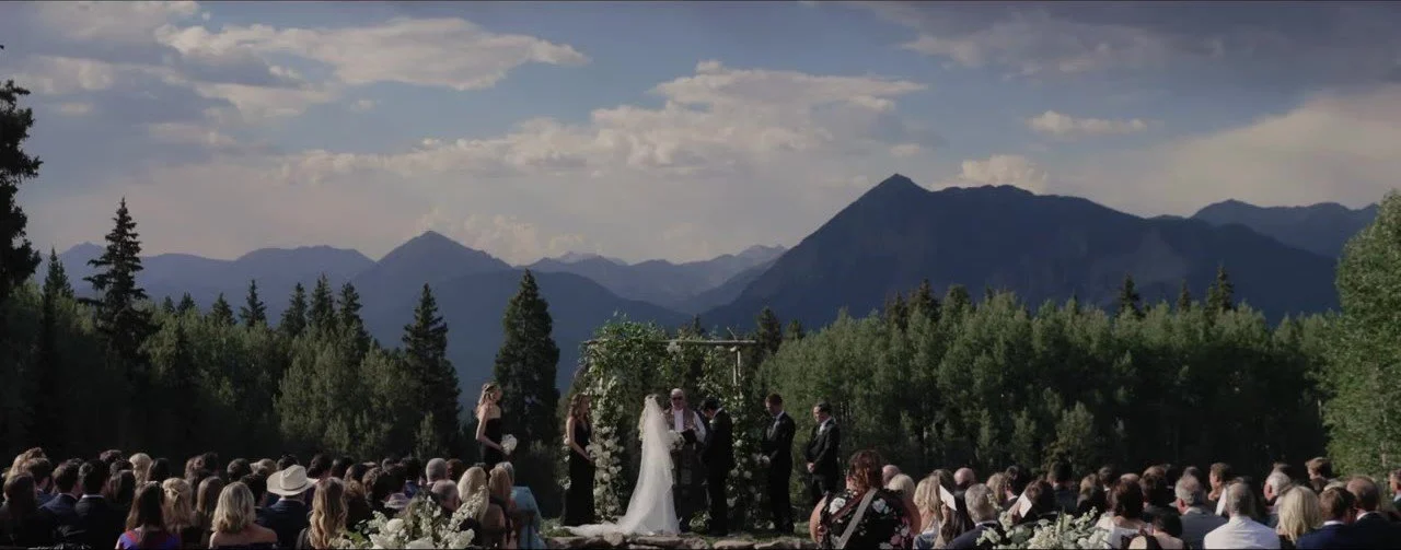 Lauren and Davey under the floral arch with guests in the foreground and Crested Butte peaks behind the trees