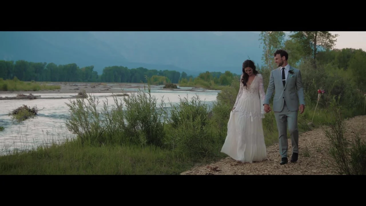 Ben and Hilah walking a riverside path in lace and a grey suit, Tetons hazy beyond the water