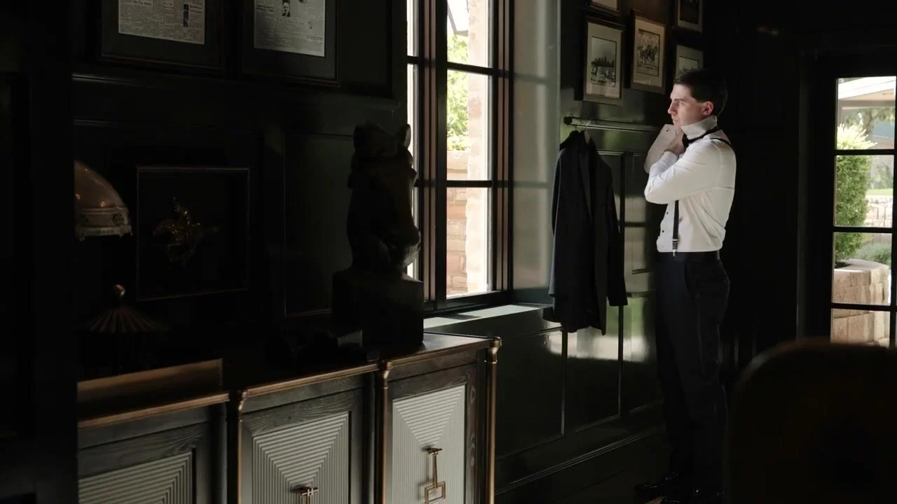 Groom in a white shirt and black suspenders adjusting his collar in a wood paneled suite at Red Berry Estate