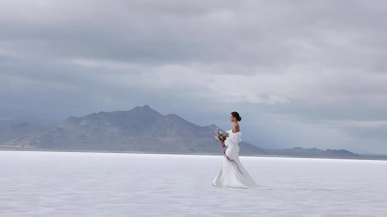 Bride standing alone on the Bonneville Salt Flats in a puffy white gown, bouquet with trailing ribbon, mountains on the horizon