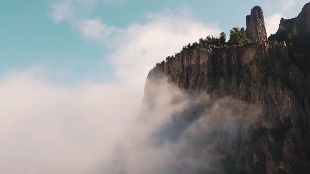 A tall rocky cliff with trees on the ridge, partly covered by white cloud on the Ouray Via Ferrata