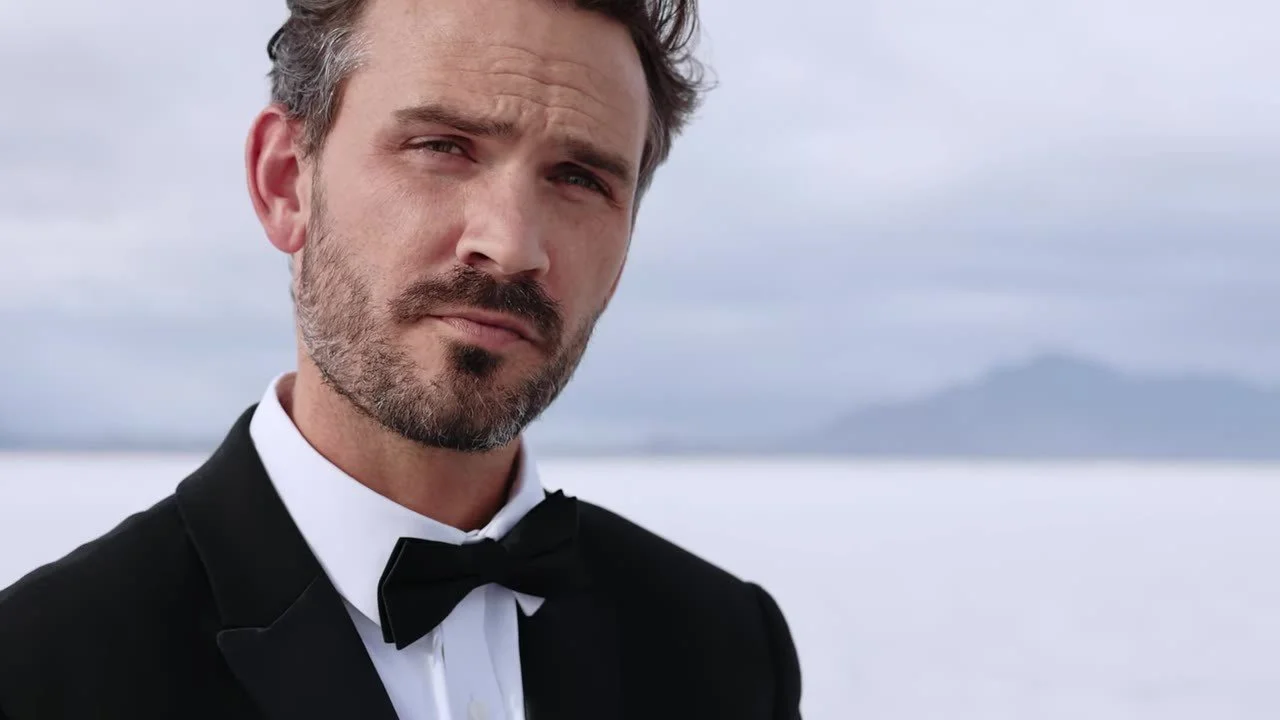 Tight portrait of the groom in a black tuxedo and bow tie on the salt flats with mountains behind him