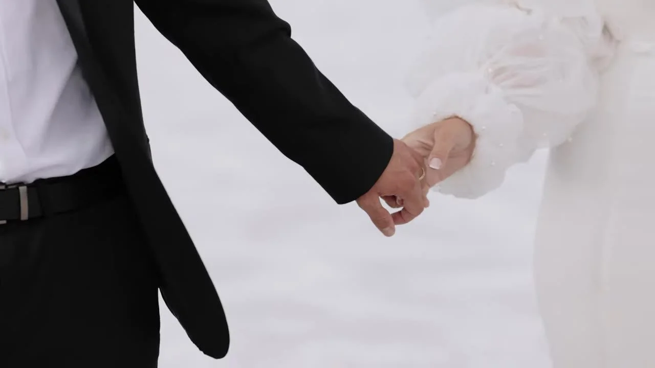 Tight shot of clasped hands, gold band, and a pearl sleeve against white salt