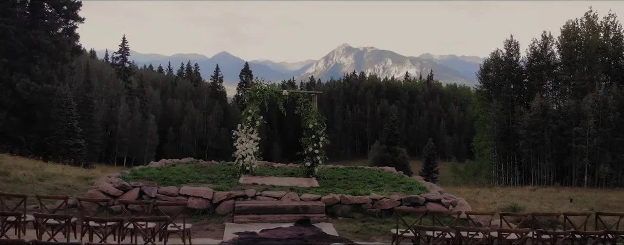 Wooden floral ceremony arch on a stone platform with Crested Butte mountains behind the trees