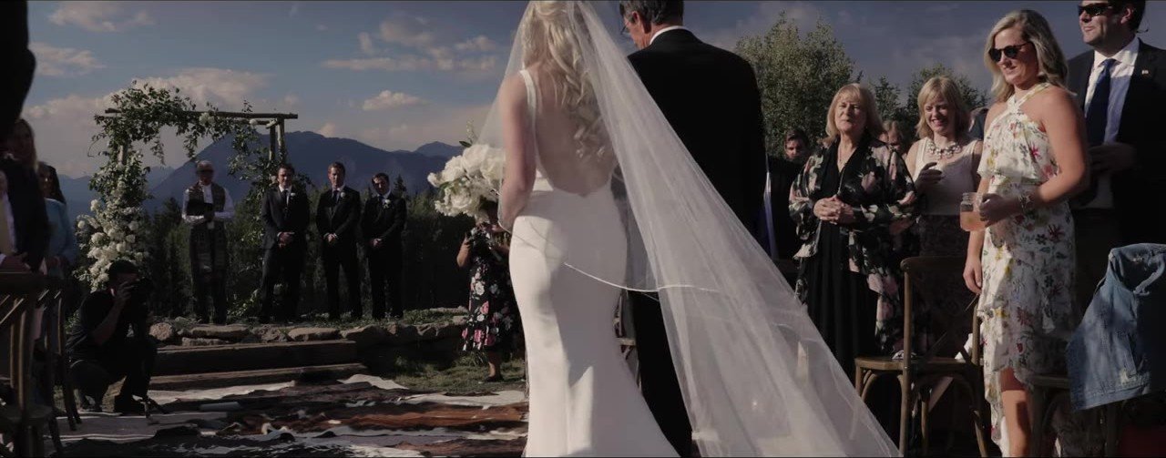 Lauren walking the aisle in a white gown and veil toward the mountain altar, guests and photographer beside her