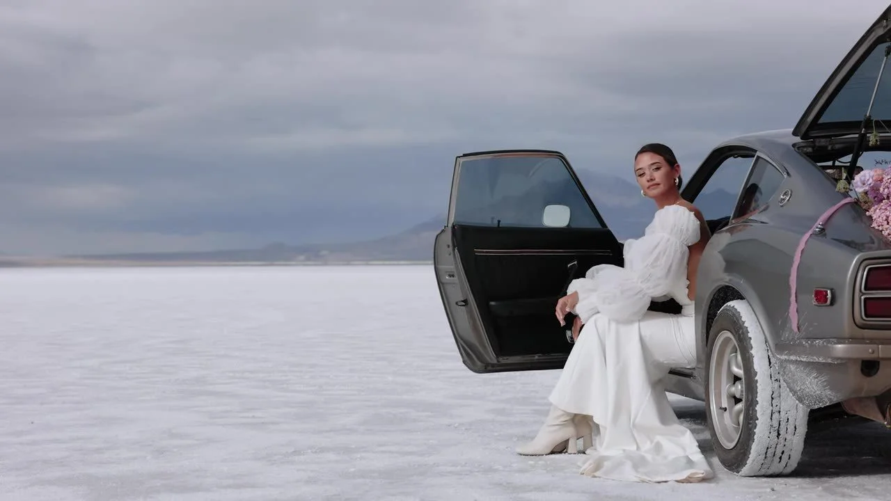 Bride in puffy sleeves and white boots sitting in the open 280Z on the salt flats, looking at camera