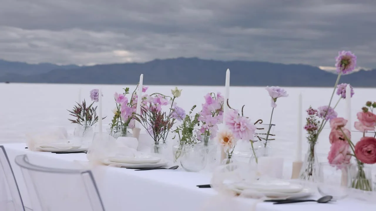 Long white table with pink flowers, taper candles, and ghost chairs set on the Bonneville Salt Flats