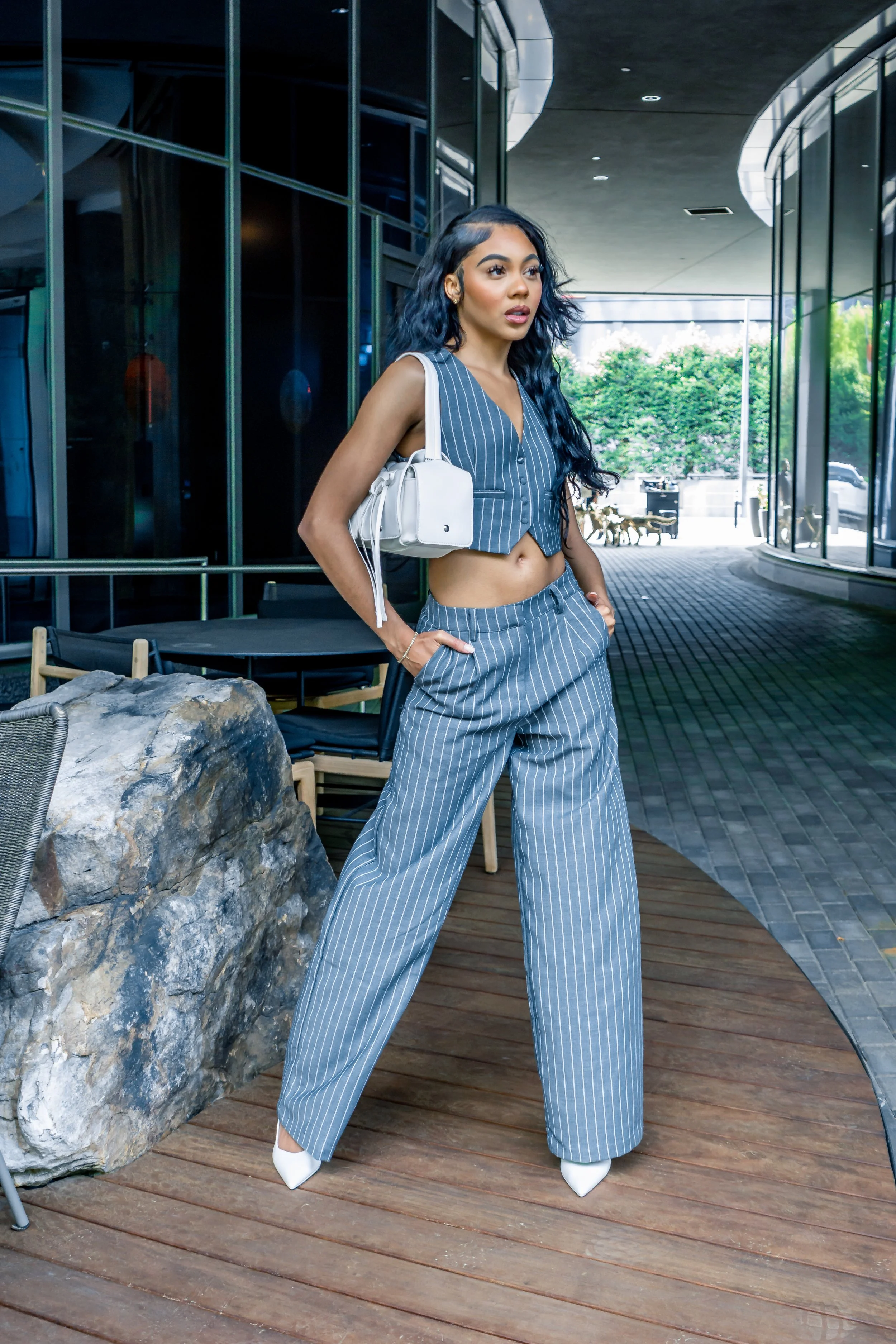 Fashion model in pinstripe suit with white purse, standing near rock, modern background.