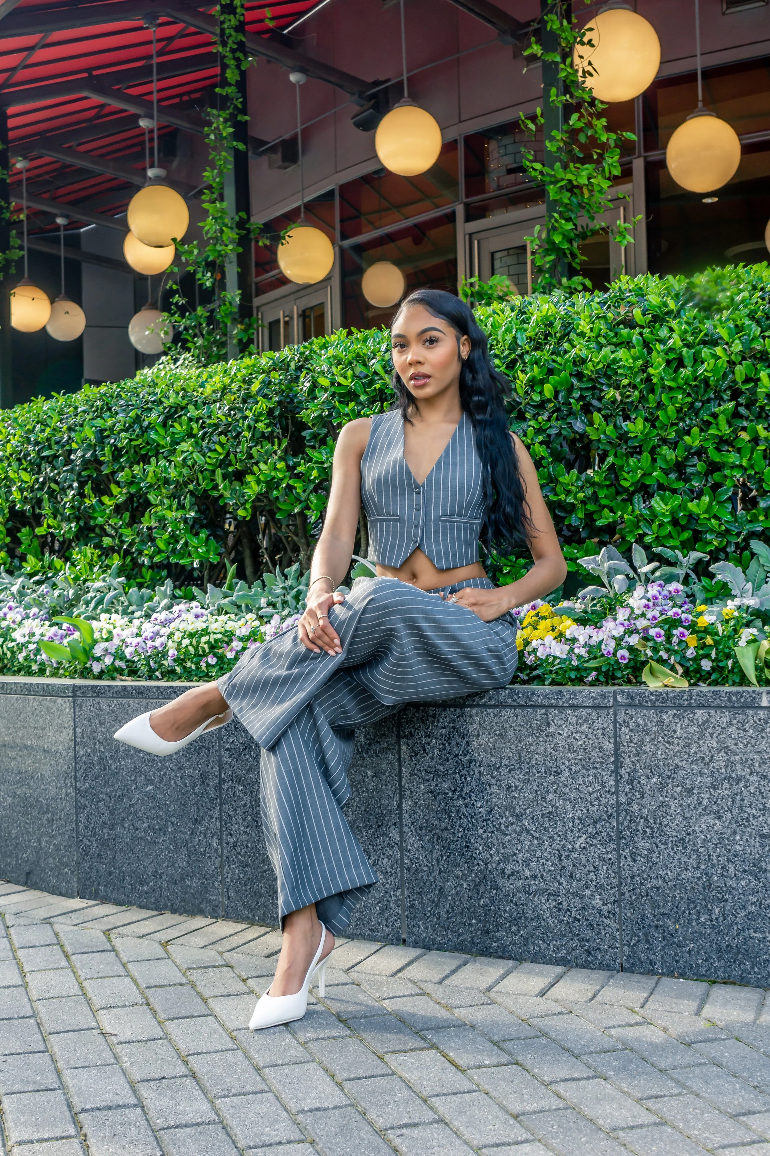 Woman in pinstripe vest and pants sitting on a ledge near greenery and flowers, wearing white heels; background features modern architecture with round hanging lamps.