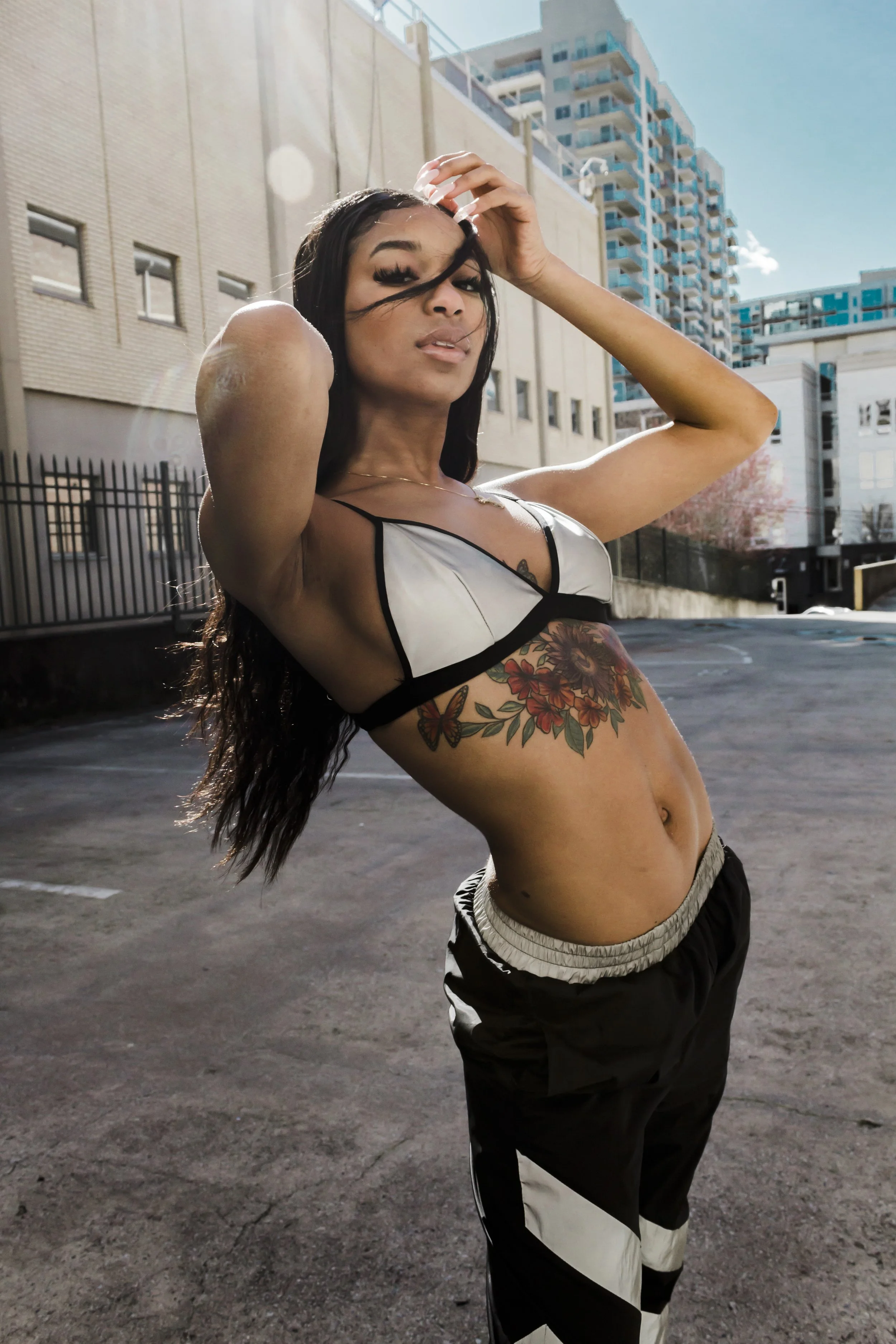 Person posing outdoors wearing a crop top and pants, with tattoos visible on their torso, in an urban setting with buildings in the background.