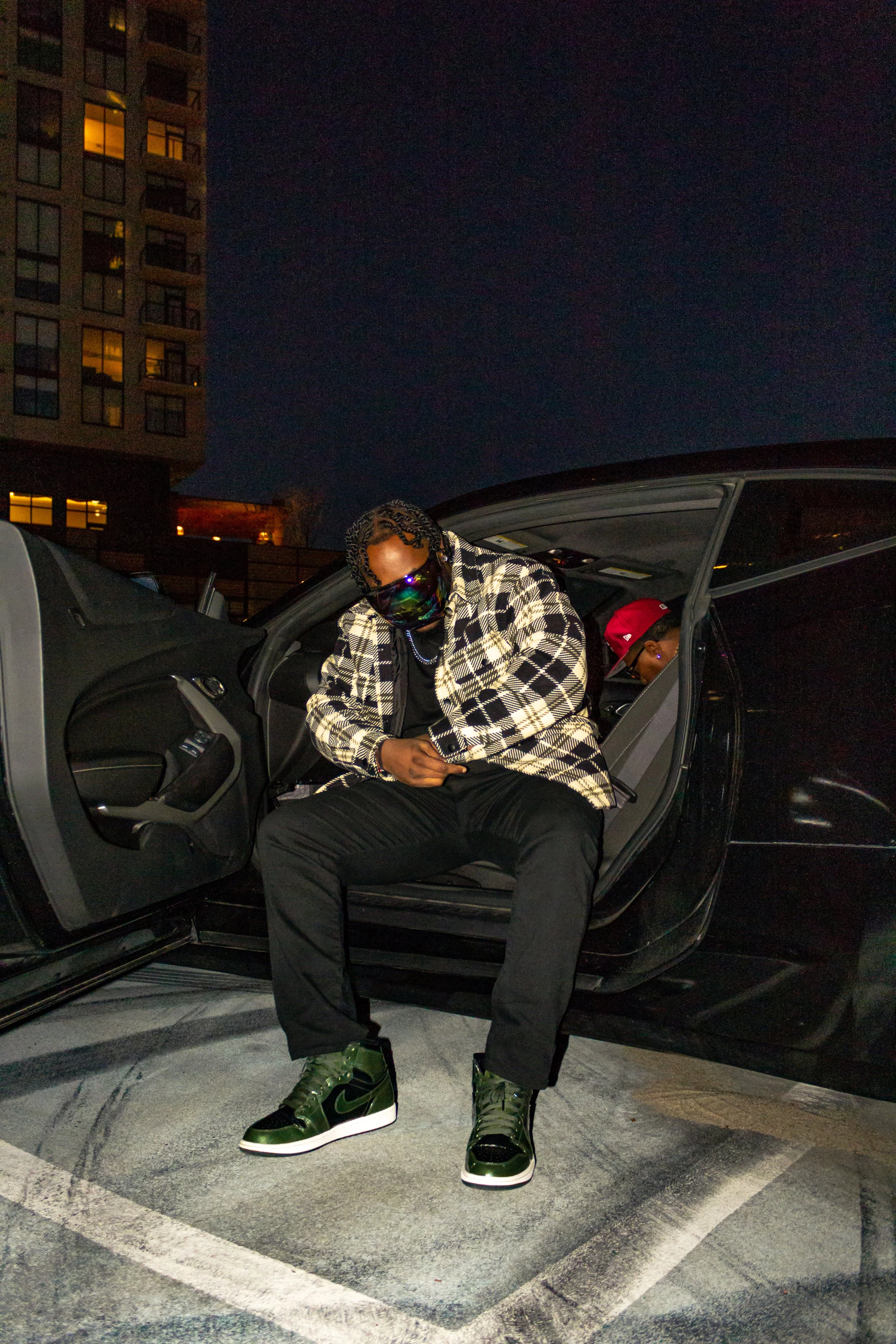 Person wearing green sneakers, black pants, and plaid jacket sitting in a car at night.