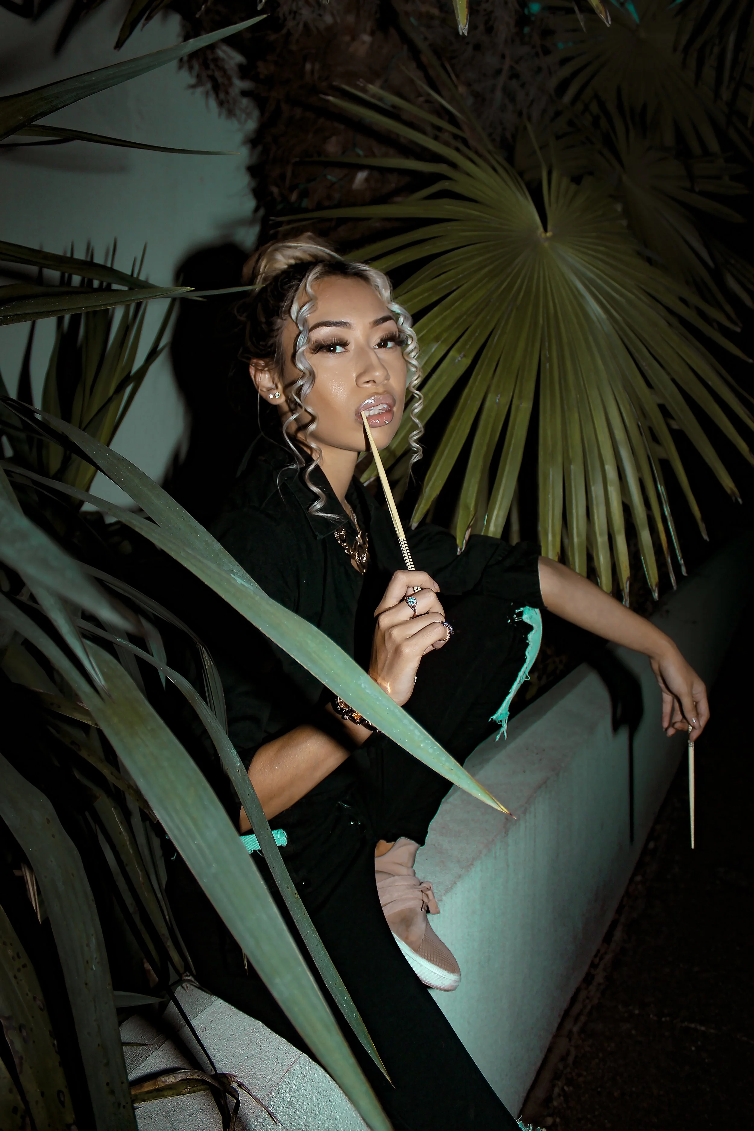 Person sitting with foliage, holding a stick, wearing black clothes and sneakers.