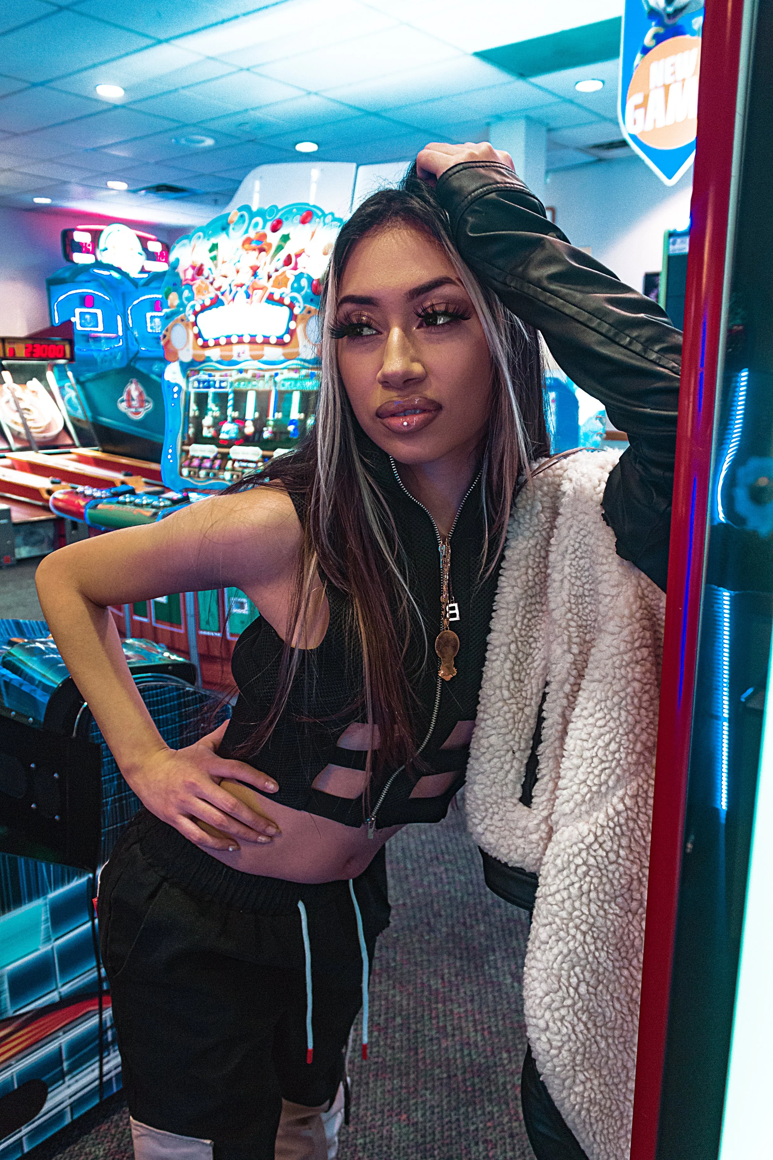 Person posing in an arcade, wearing a stylish outfit, with gaming machines and neon lights in the background.
