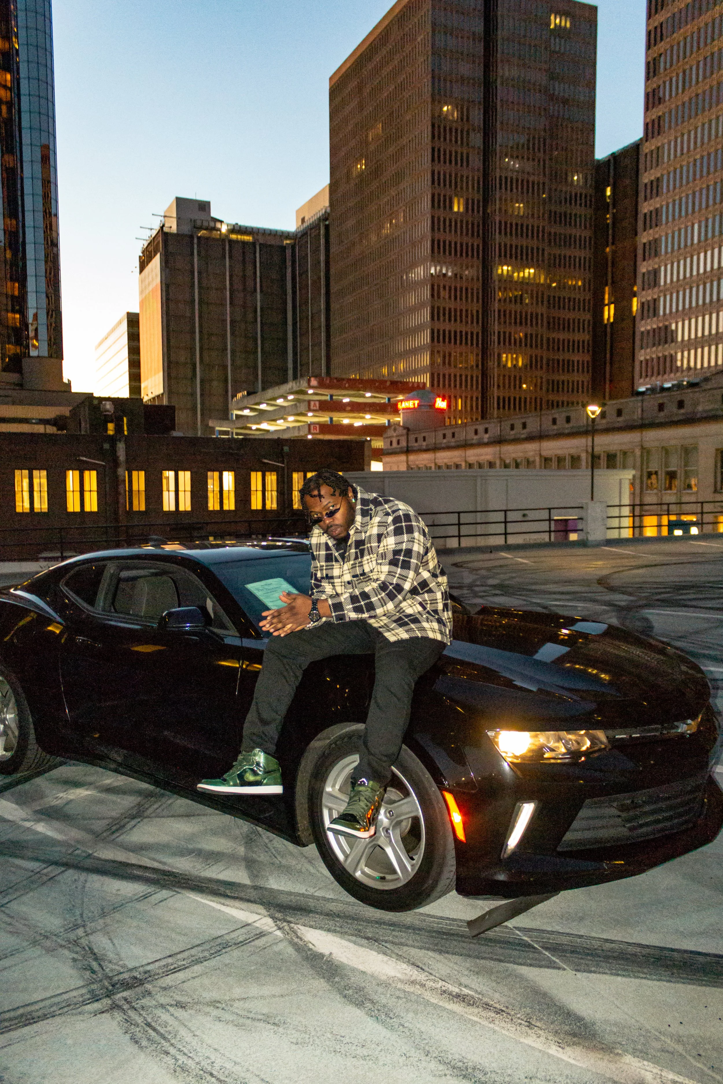 Person sitting on a black car in an urban cityscape background with tall buildings during dusk.