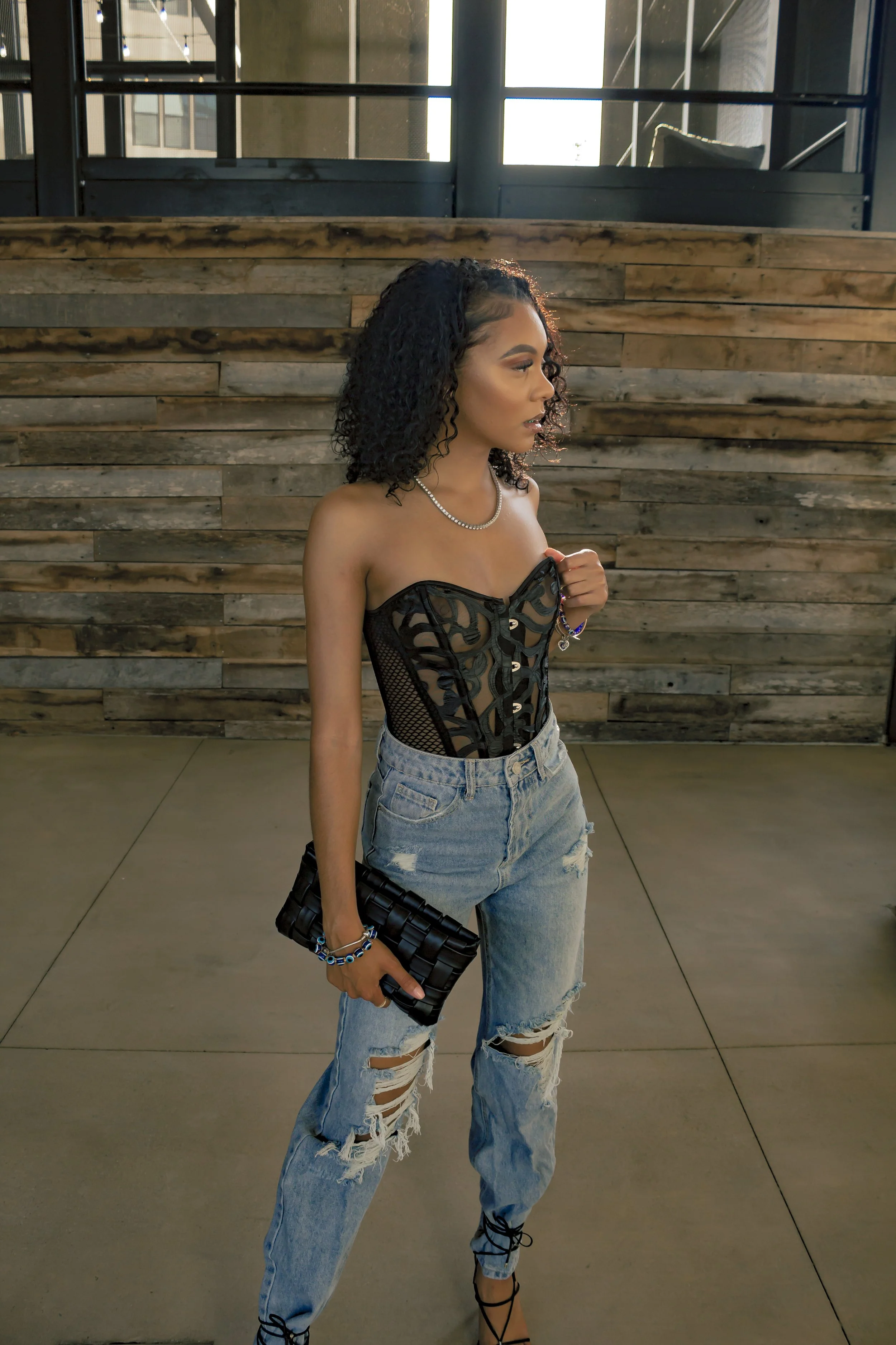 Woman in a black corset and ripped jeans holding a clutch against a wooden wall.