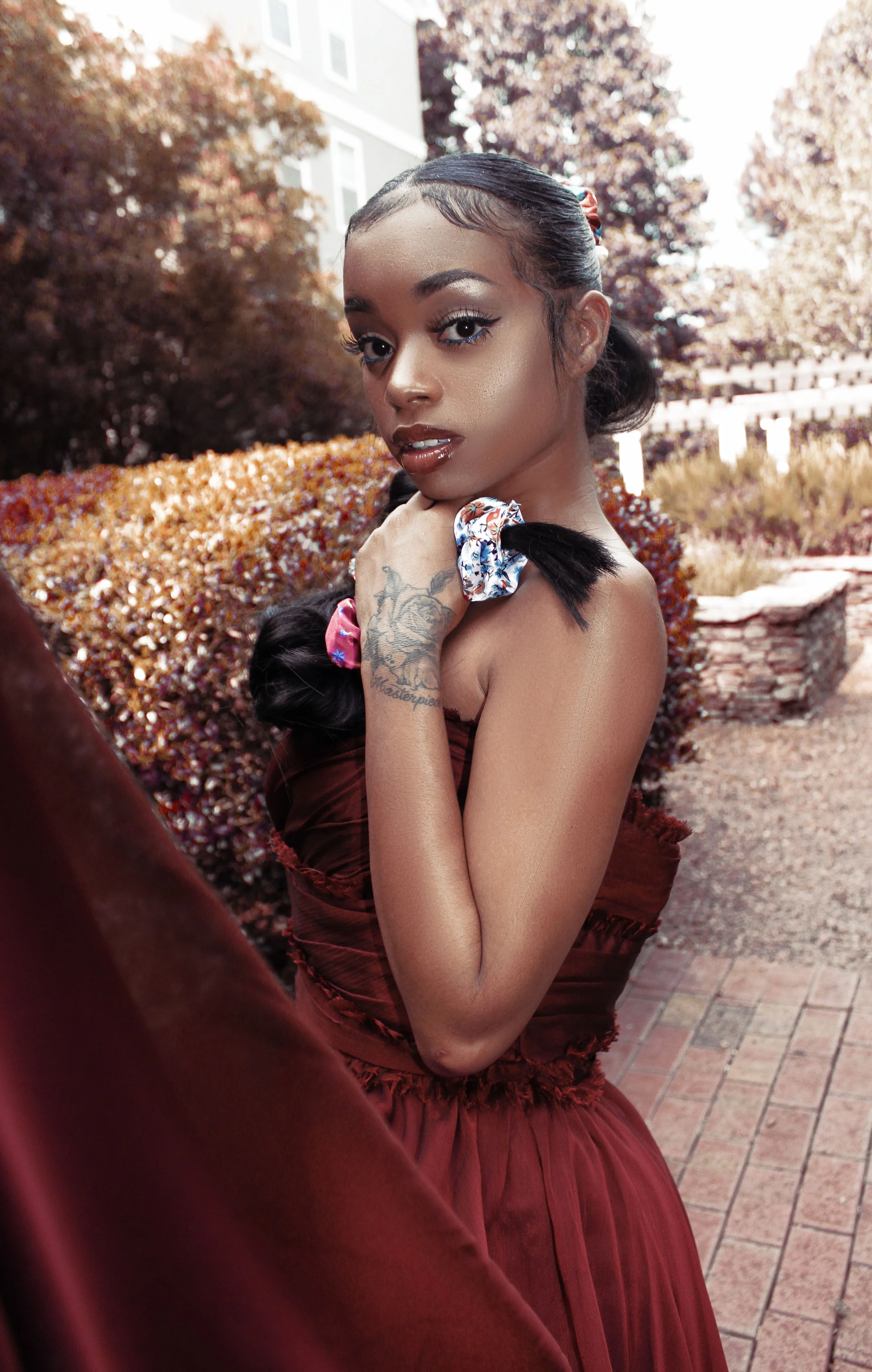 Woman in a red dress holding a floral-patterned hair tie, standing outdoors with trees in the background.