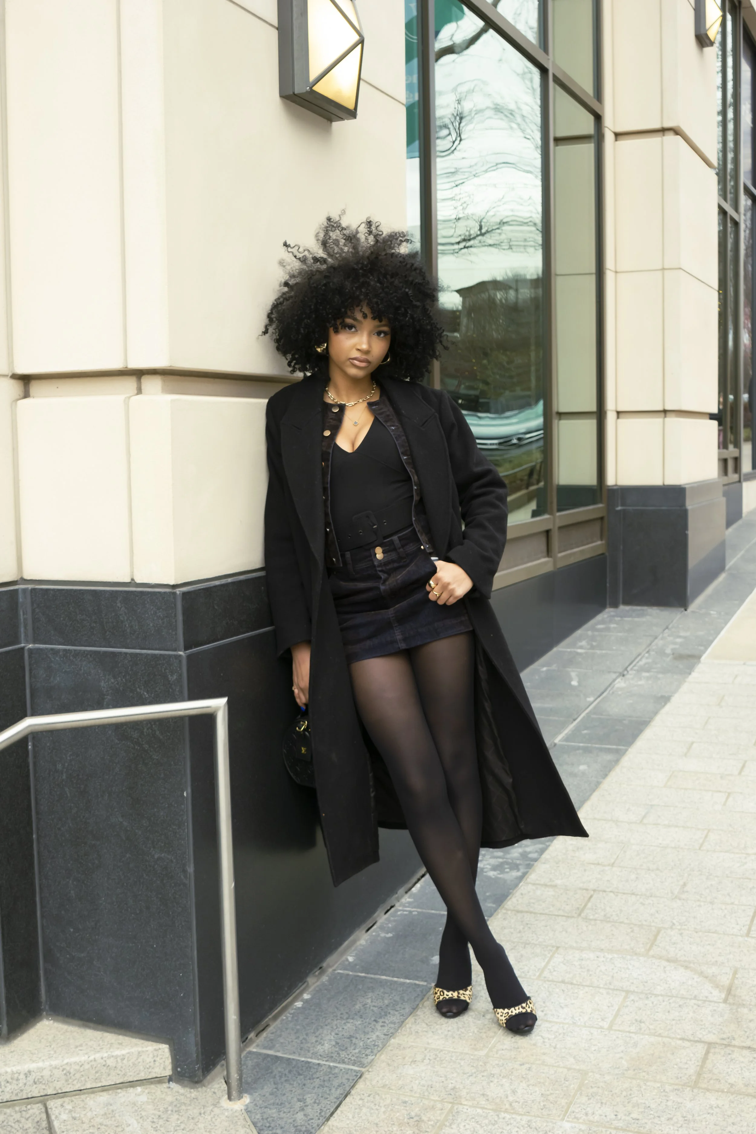 Fashionable woman in black outfit with afro hairstyle leaning against a building wall.