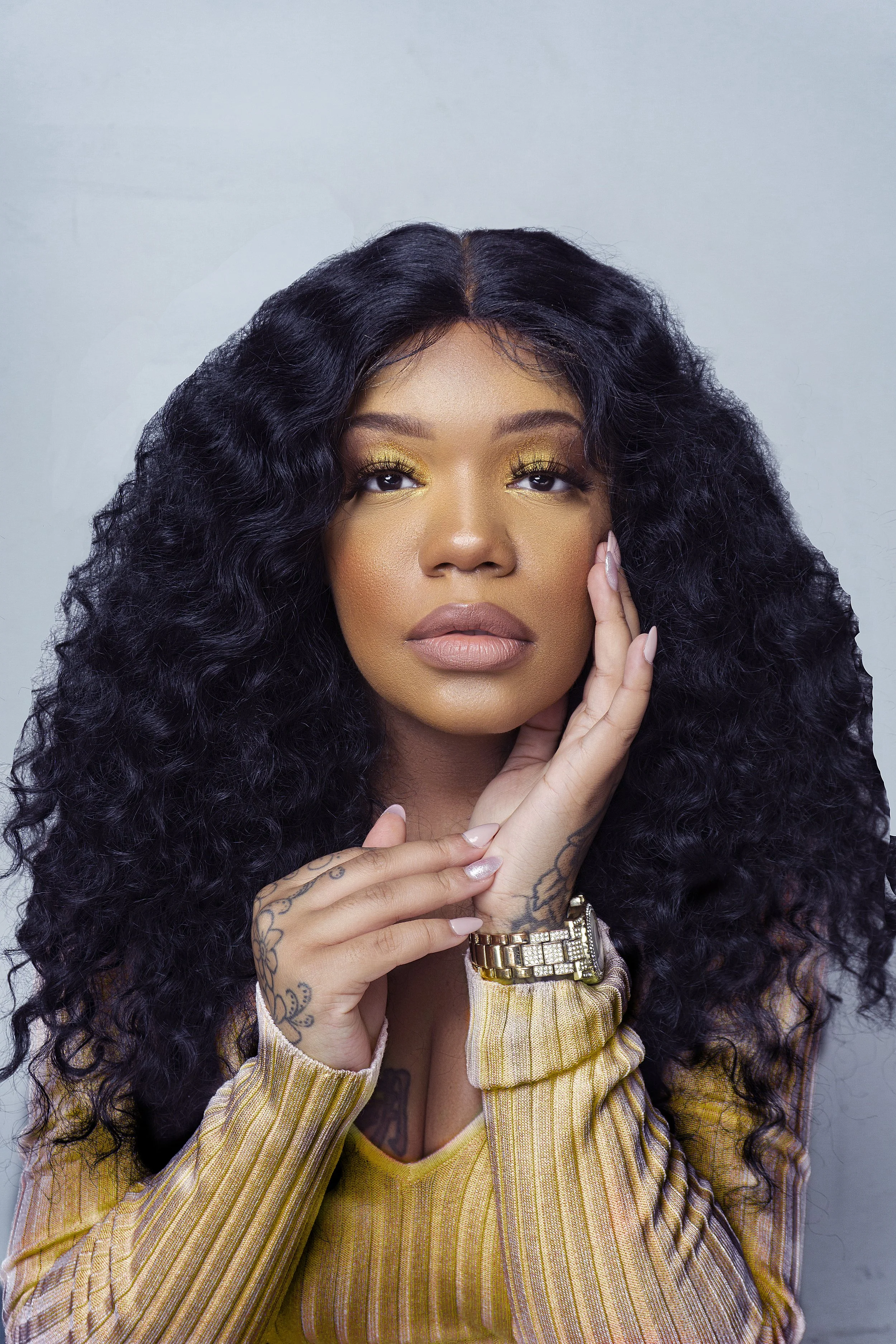 Woman with curly hair, yellow top, tattoos, gold watch