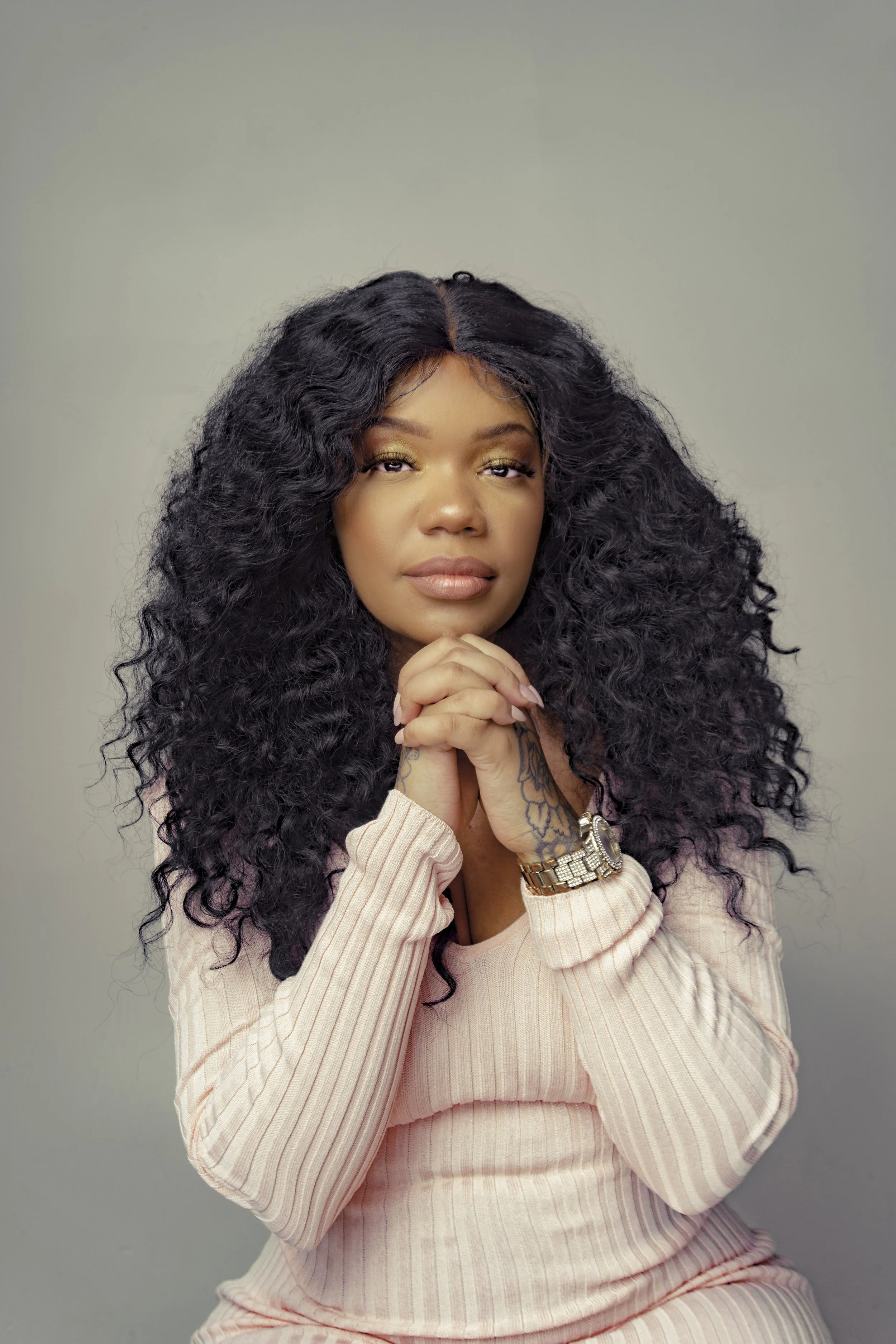 Woman with long curly hair, wearing a pink ribbed sweater, hands clasped together, looking forward.