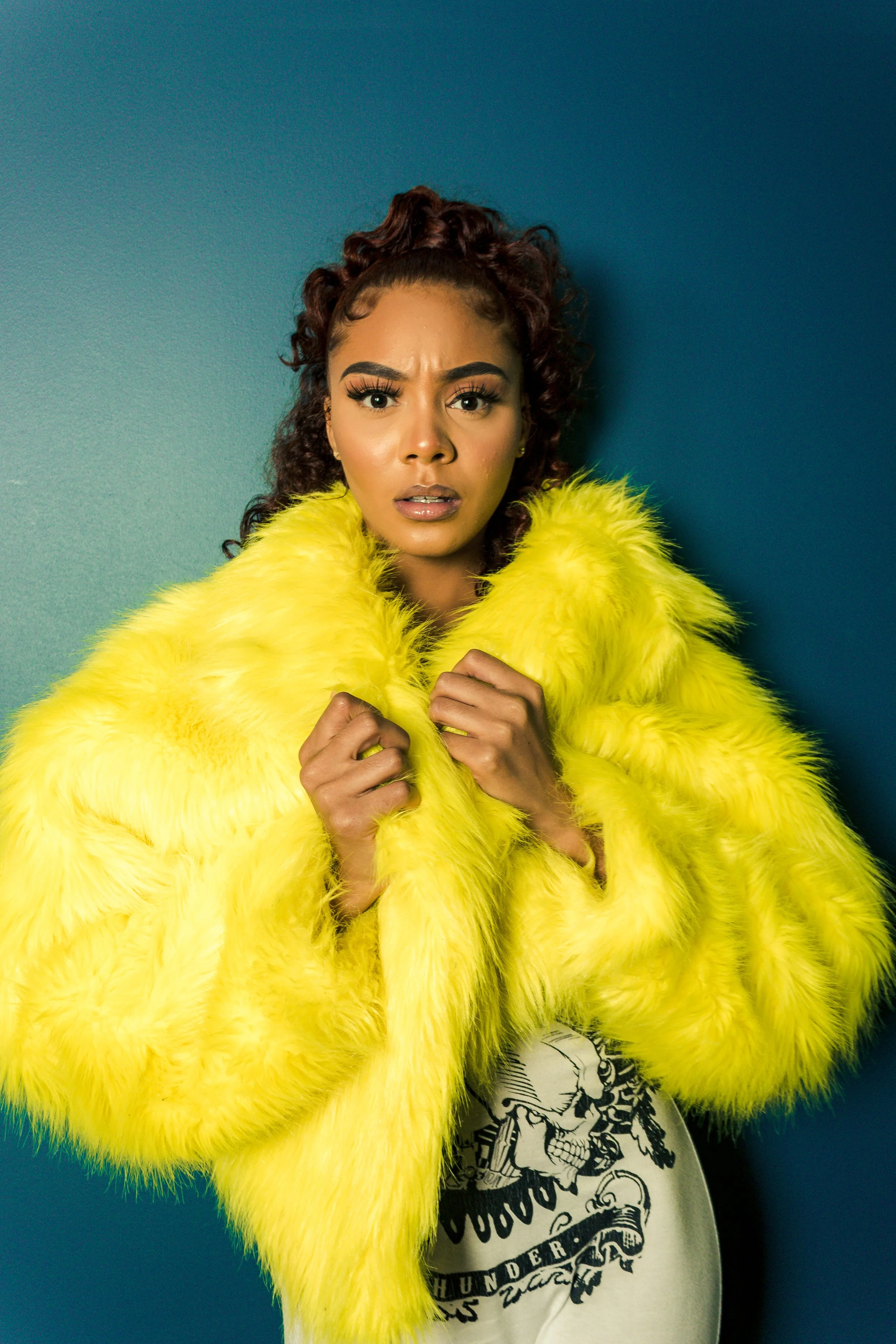 Person wearing a yellow faux fur coat and graphic t-shirt against a blue background.