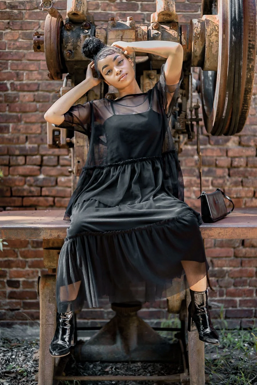 Person in black dress sitting on industrial machinery against brick wall.