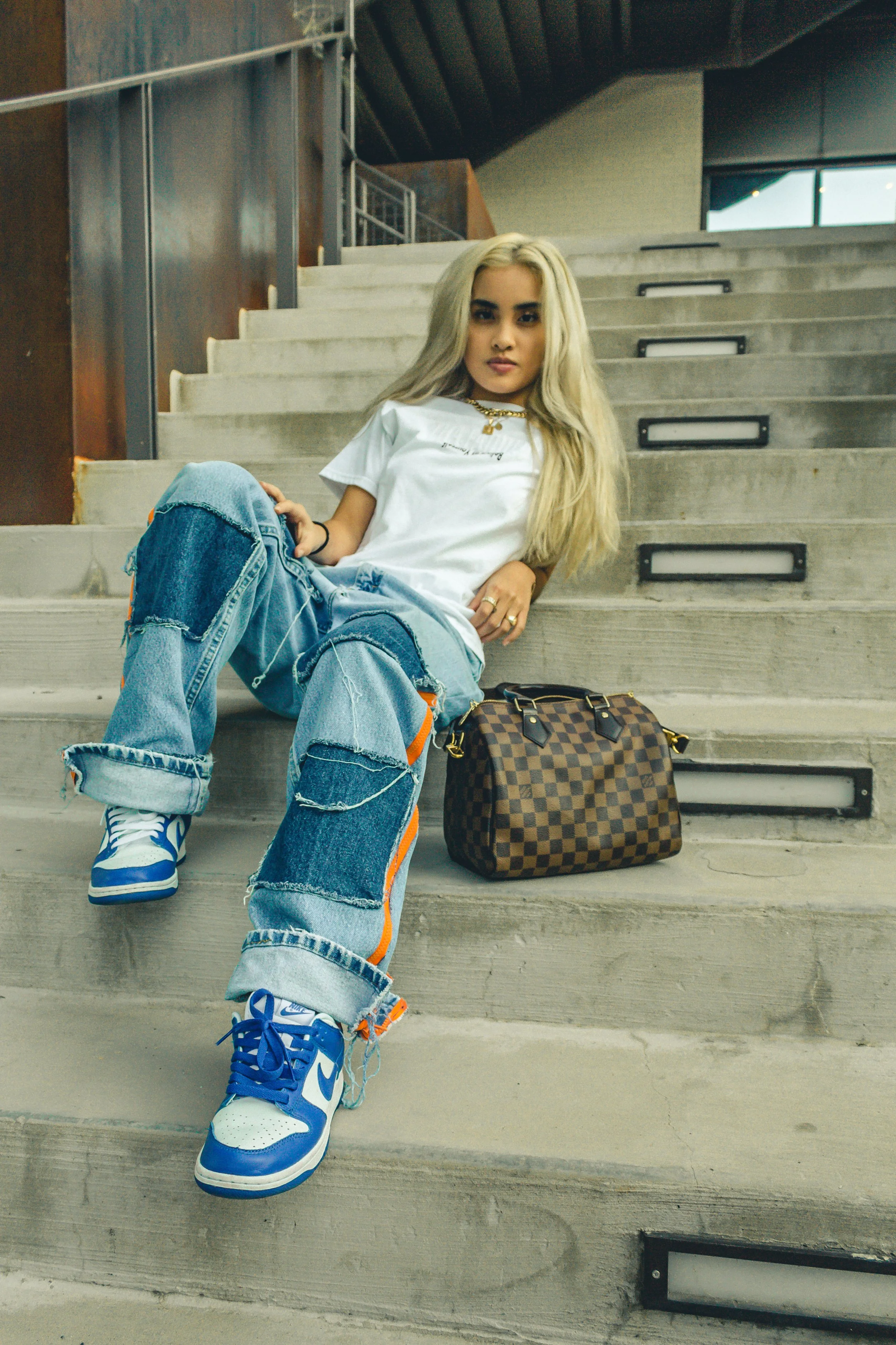 Person sitting on stairs, wearing blue patchwork jeans, white t-shirt, and blue sneakers, with a brown checkered handbag beside them.