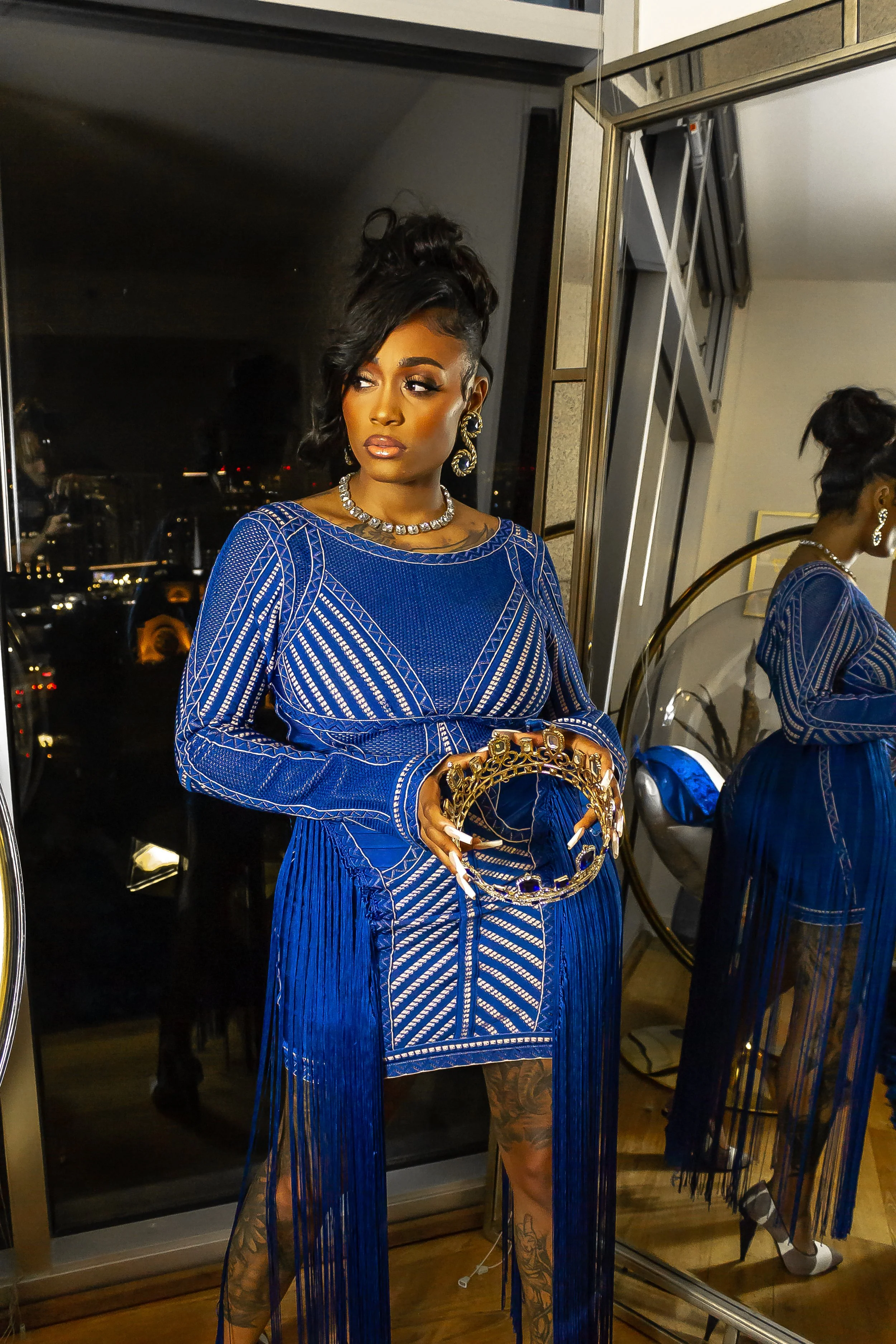 Woman in blue dress holding a crown, standing by a window with city lights visible.