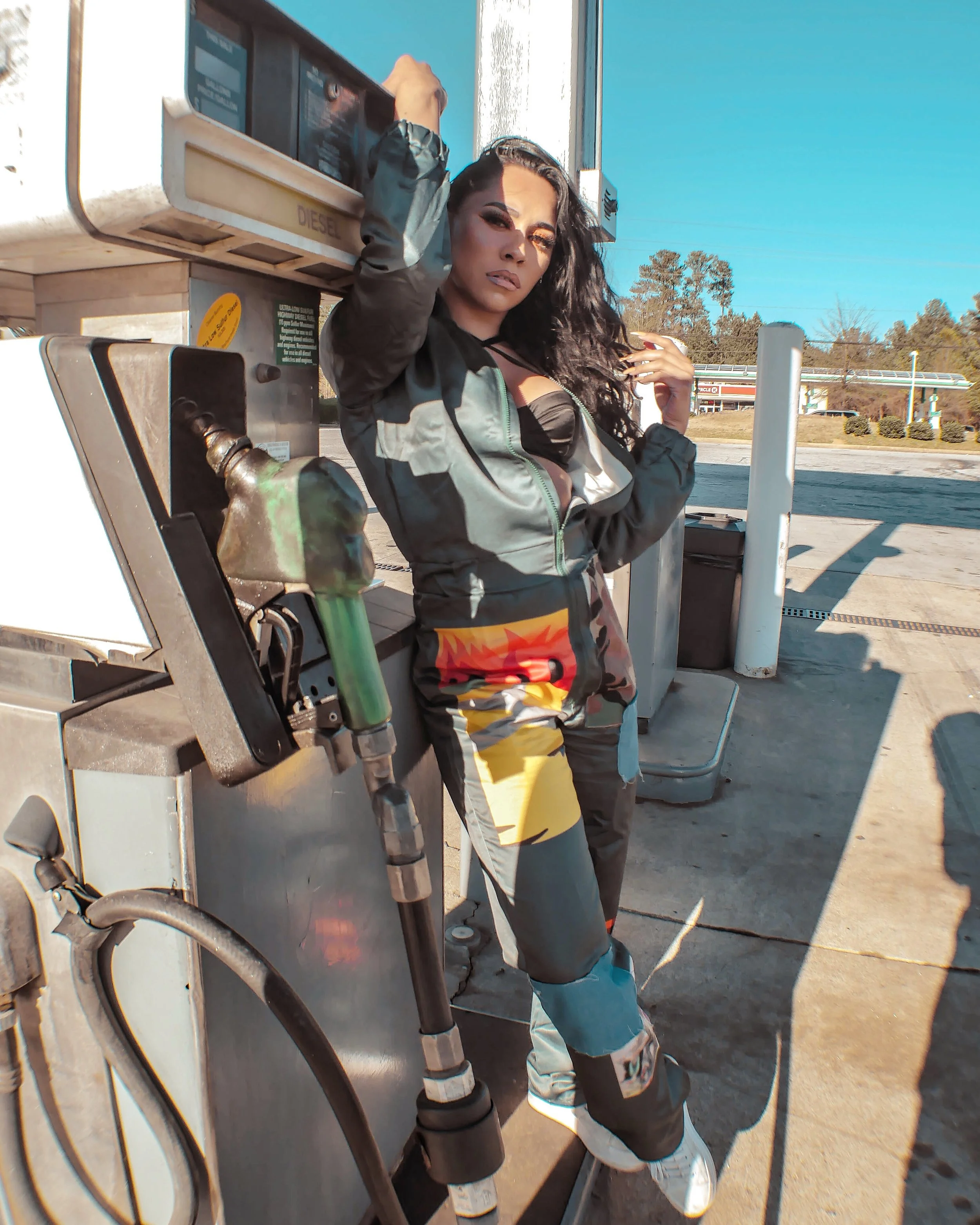 Person posing next to a fuel pump at a gas station, wearing a colorful outfit and white sneakers, with trees and a clear sky in the background.
