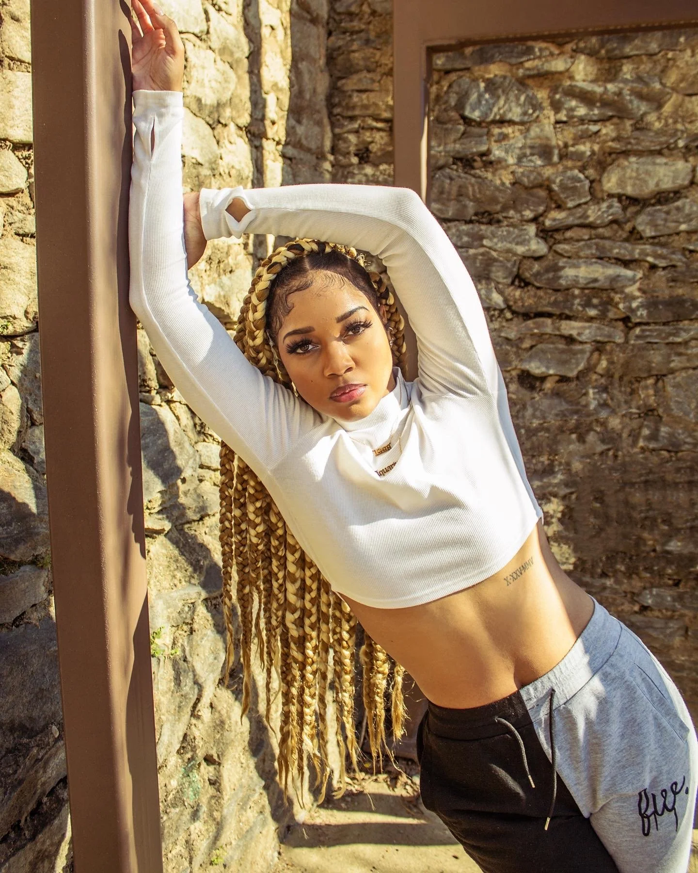Person with long braids leaning against stone wall, wearing a white crop top and gray sweatpants.